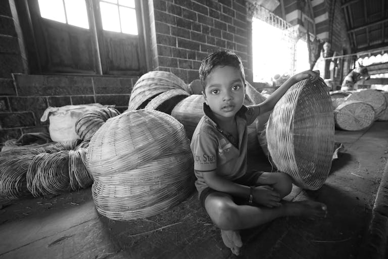 Black and white photo of a child with woven baskets indoors, highlighting traditional craftsmanship.