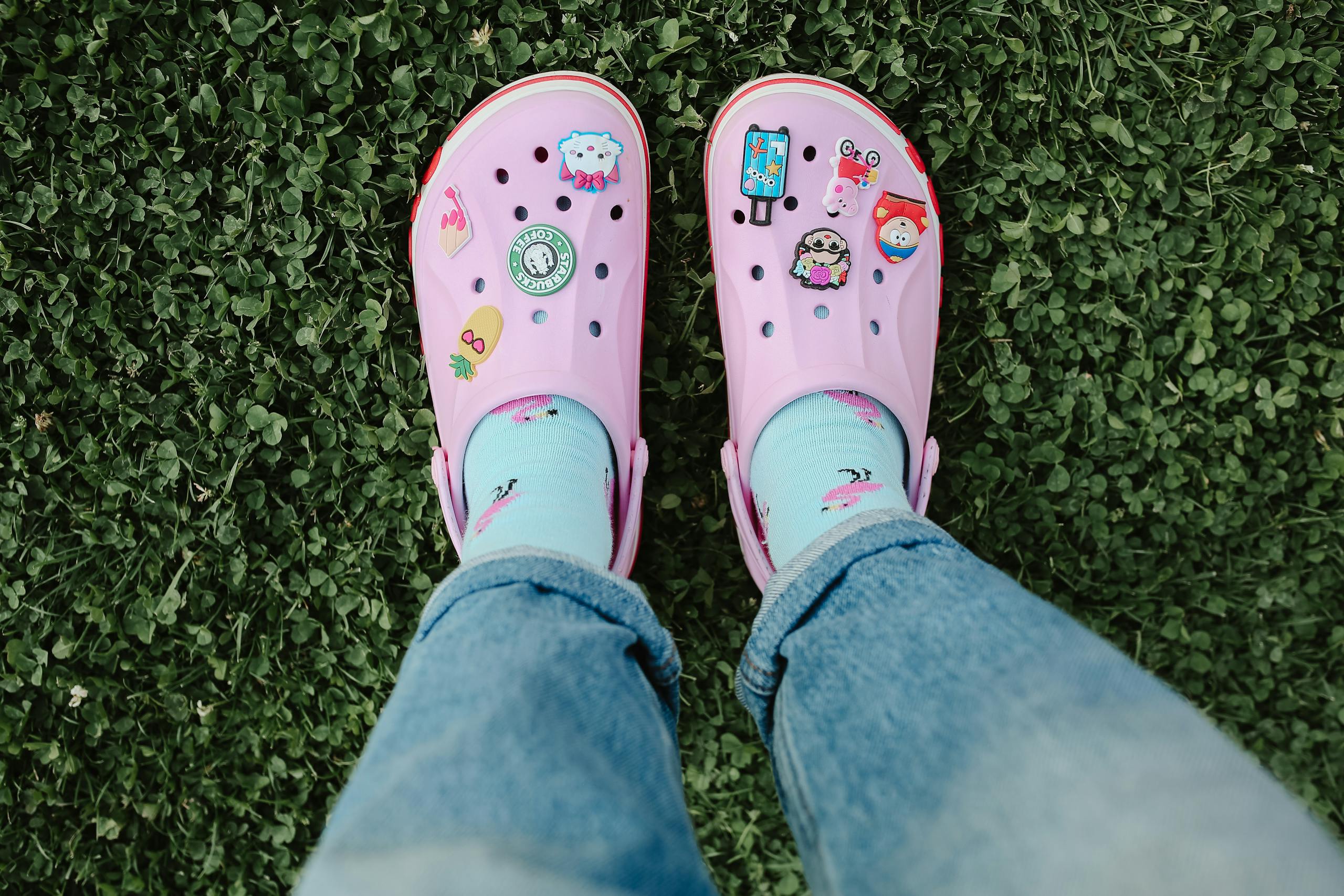 Bright pink crocs adorned with stickers, worn with blue socks, on lush green grass.