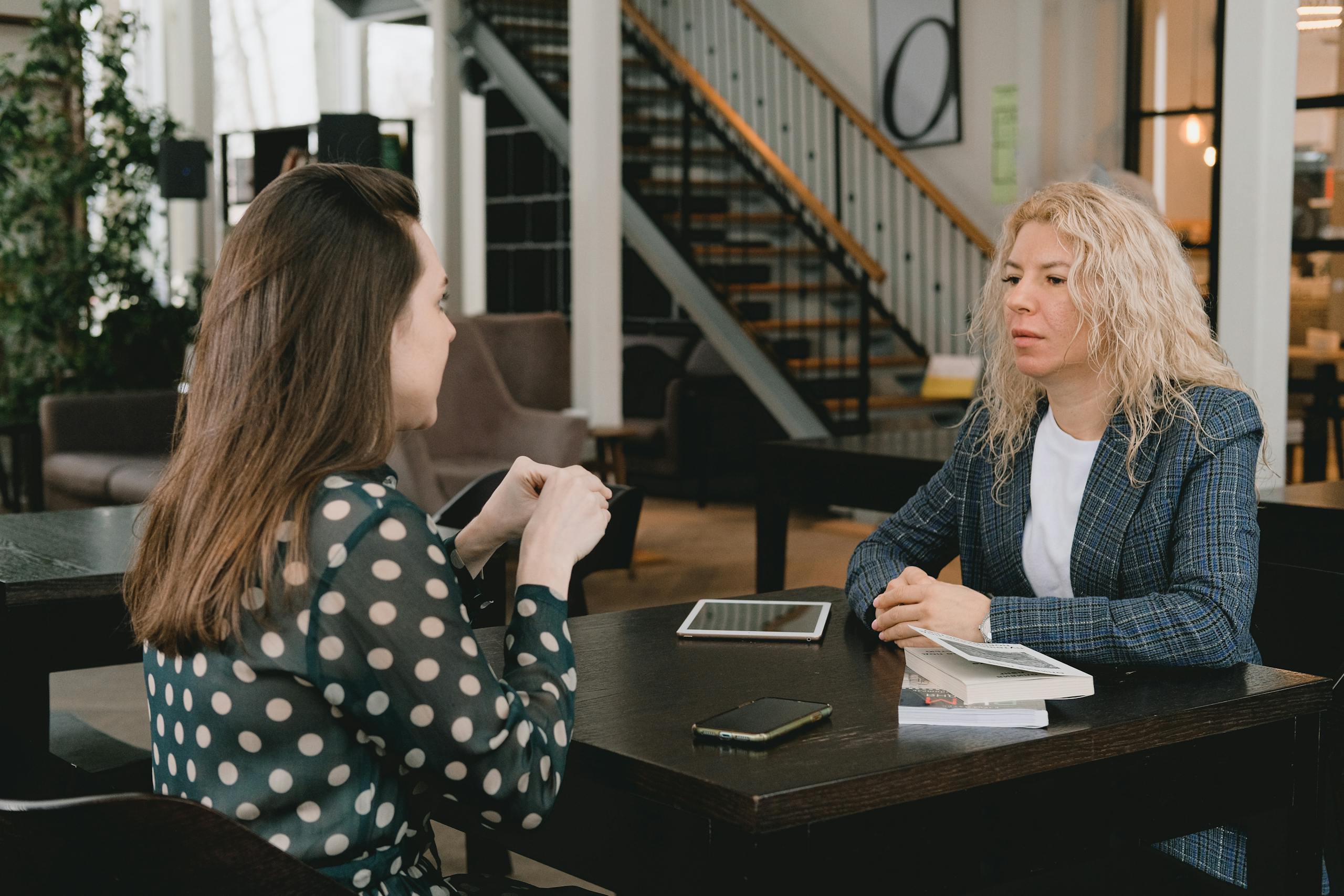 Businesswomen sitting at table with book and devices and having conversation about project in modern workplace