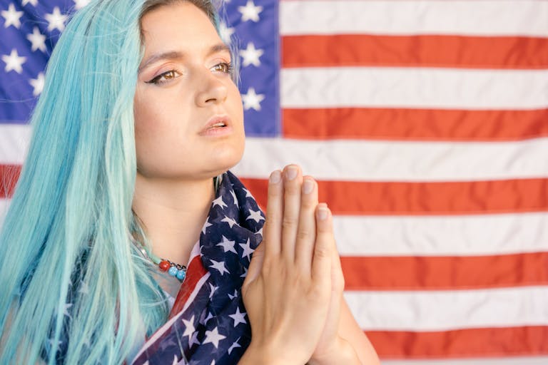 Caucasian woman with blue hair in front of American flag, hands in prayer.