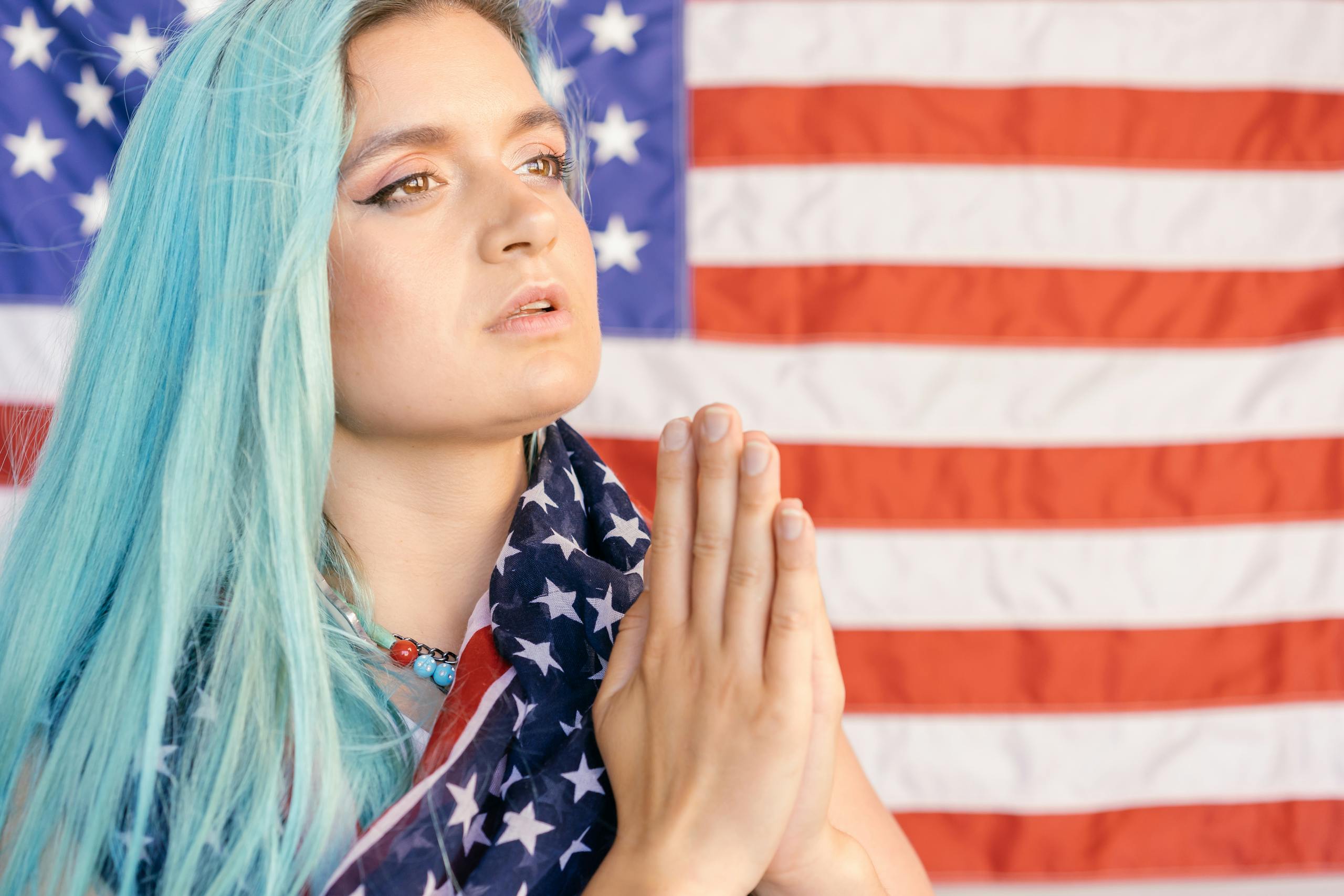 Caucasian woman with blue hair in front of American flag, hands in prayer.