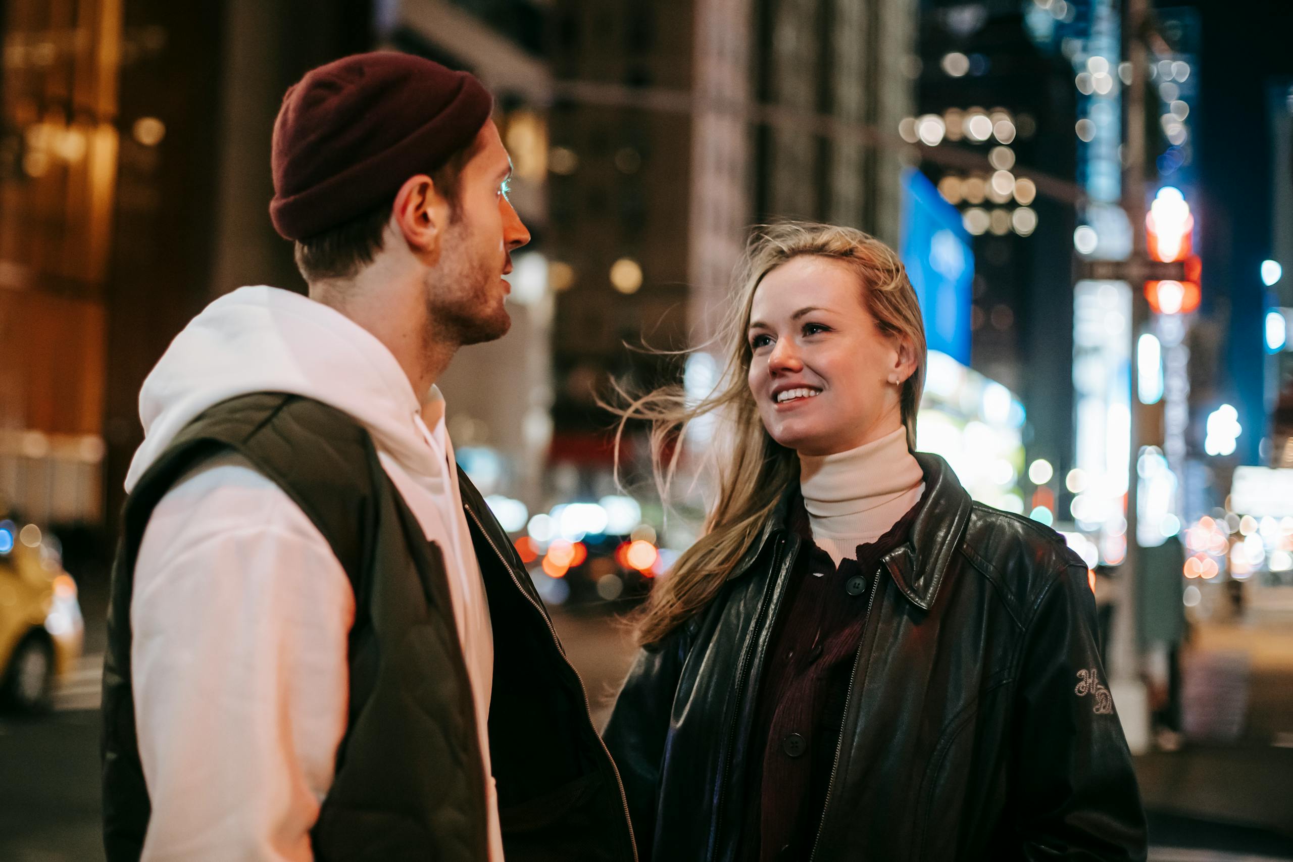 Cheerful couple in warm wear standing near road with cars on street with modern buildings at night time on blurred background