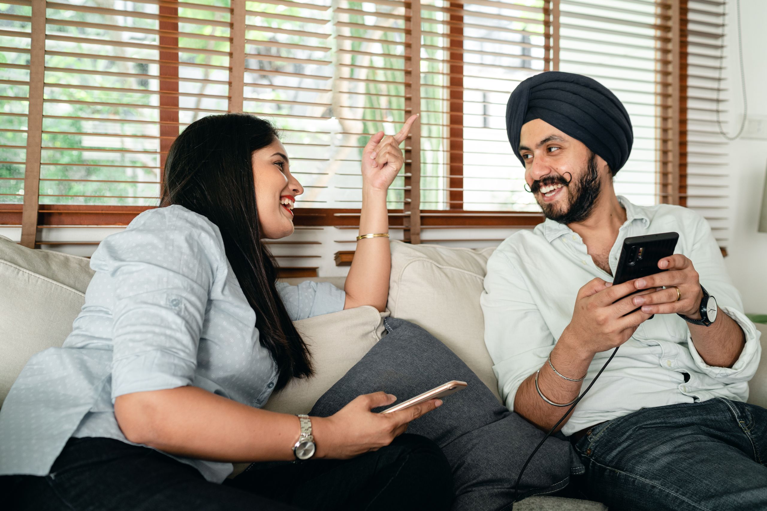 Cheerful young Indian couple in casual outfits with smartphone laughing and looking at each other on comfortable sofa during weekend
