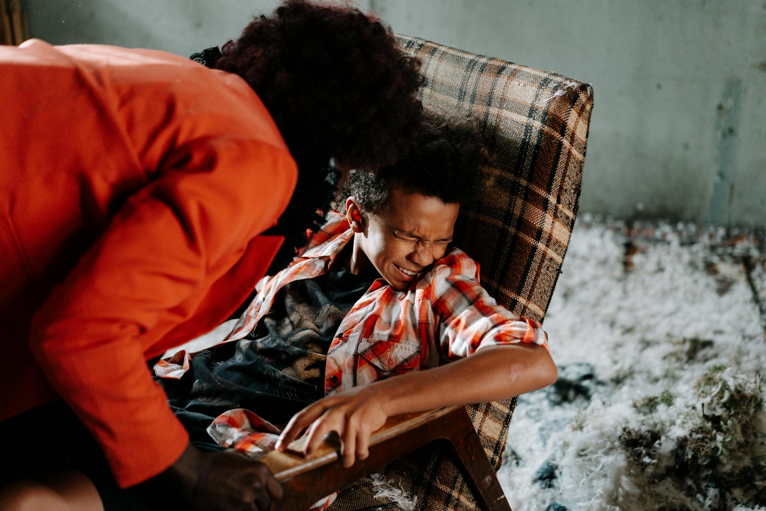 Child in distress on checkered chair with adult leaning over ominously inside a dimly lit room.