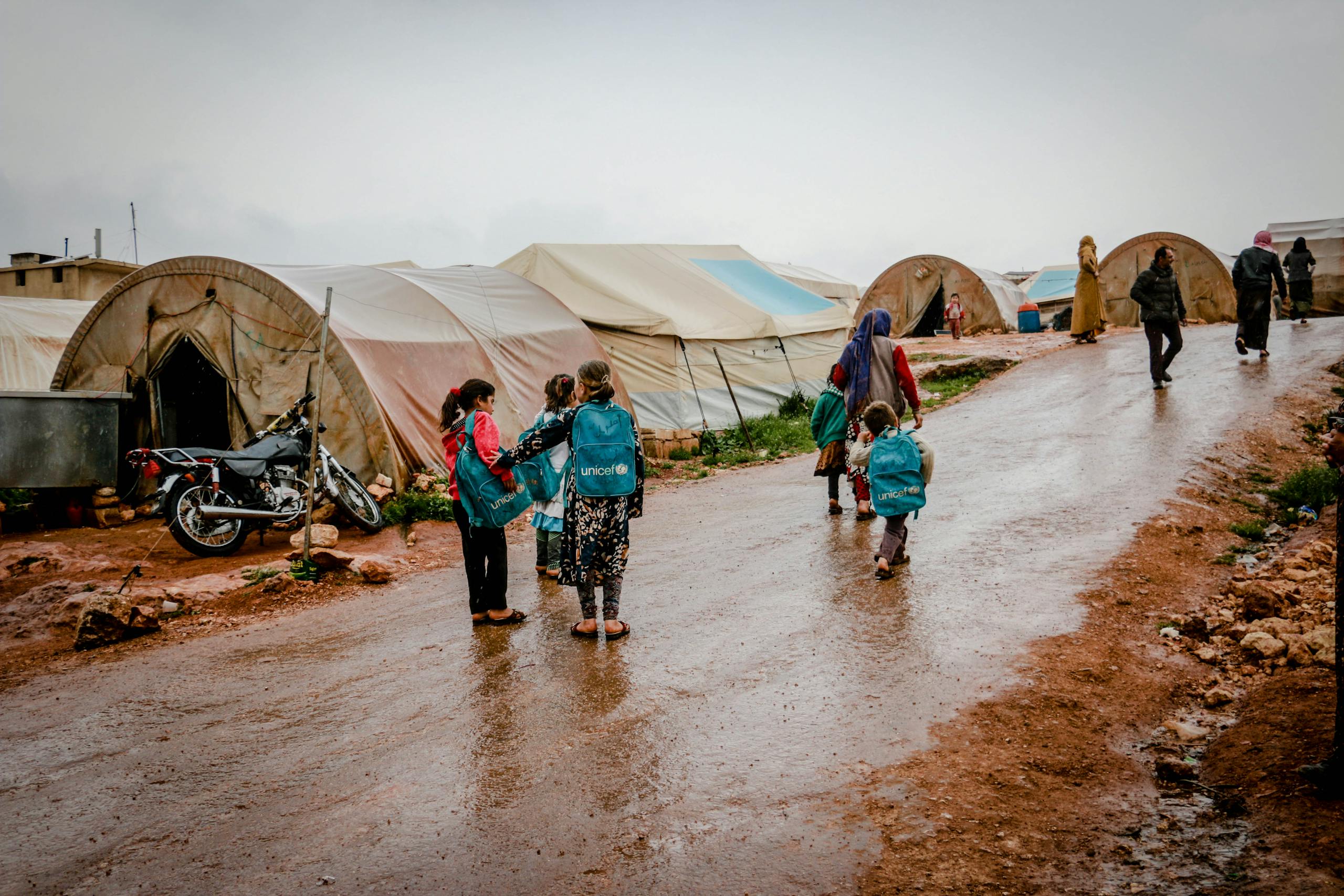 Children with backpacks walking in a rainy refugee camp in Idlib, Syria, capturing resilience and hope.
