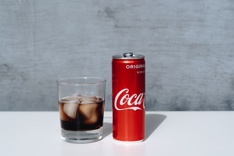 Chilled Coca-Cola can and drink with ice cubes in glass on minimalistic surface.