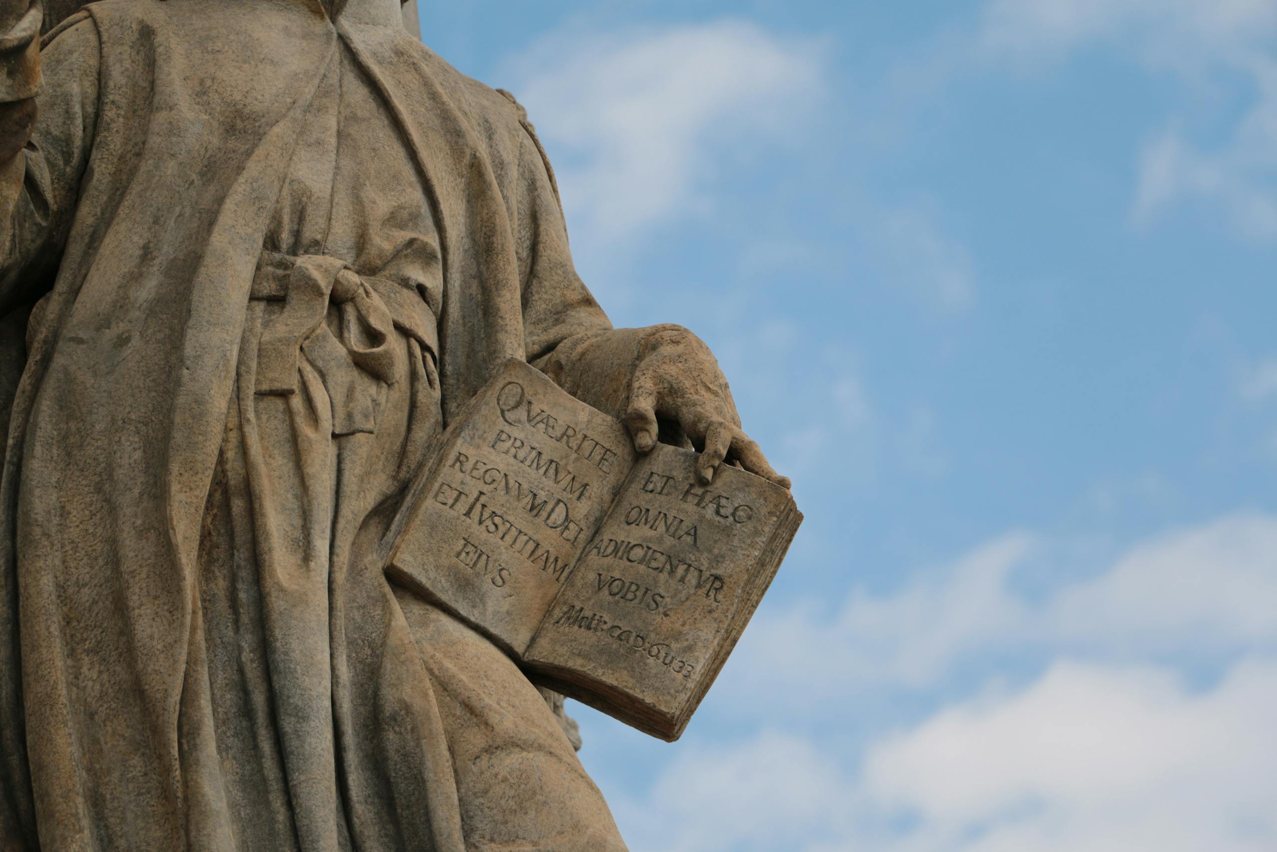 Close-up of a historical stone statue holding an open book against blue sky.