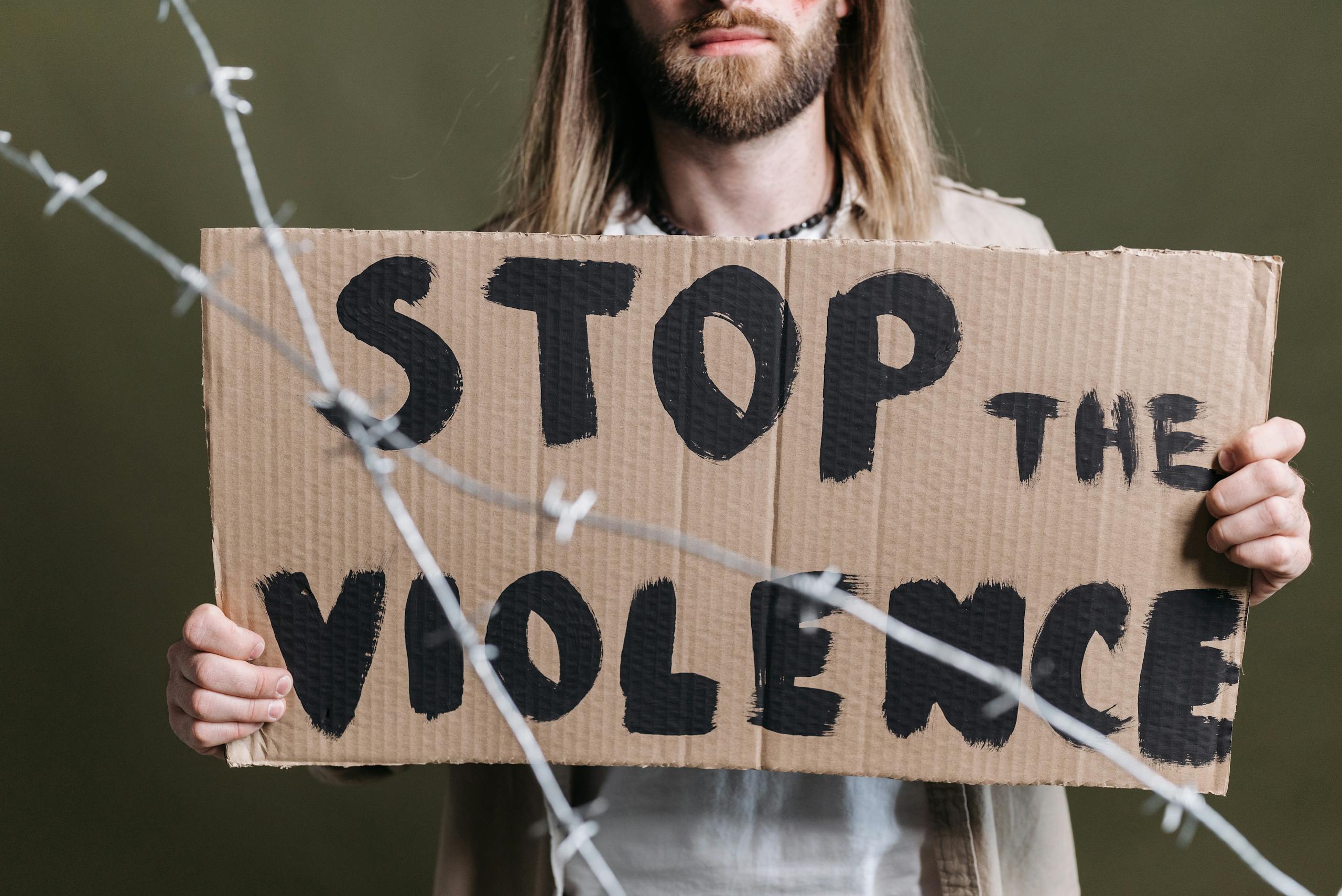 Close-up of a protester holding a cardboard sign reading 'Stop the Violence' with barb wire in focus.