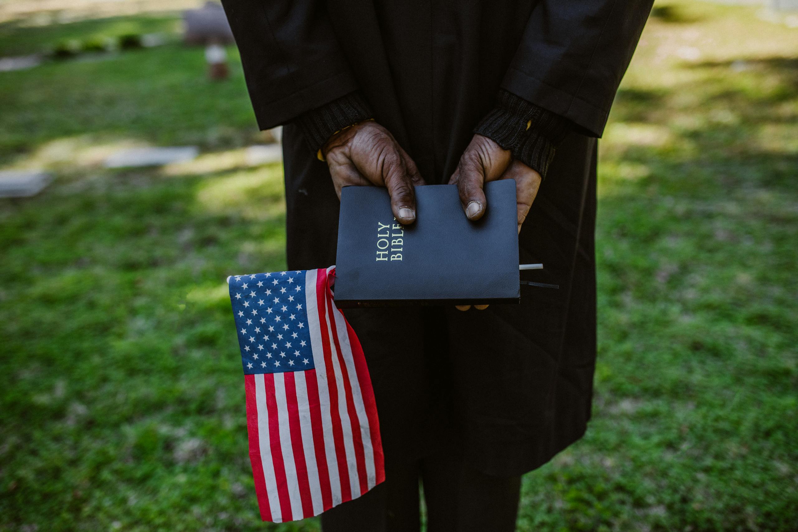 Close-up of hands holding a Holy Bible and American flag in a green outdoor setting.