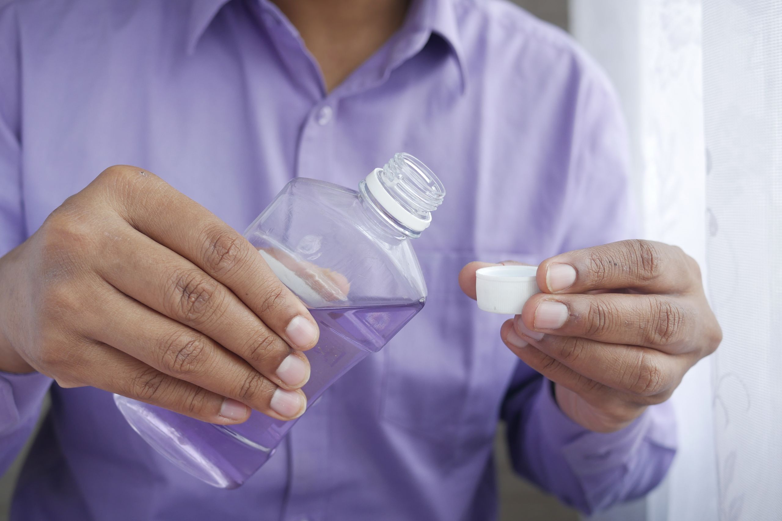 Close-up of hands holding a purple plastic bottle while pouring mouthwash into a cap.