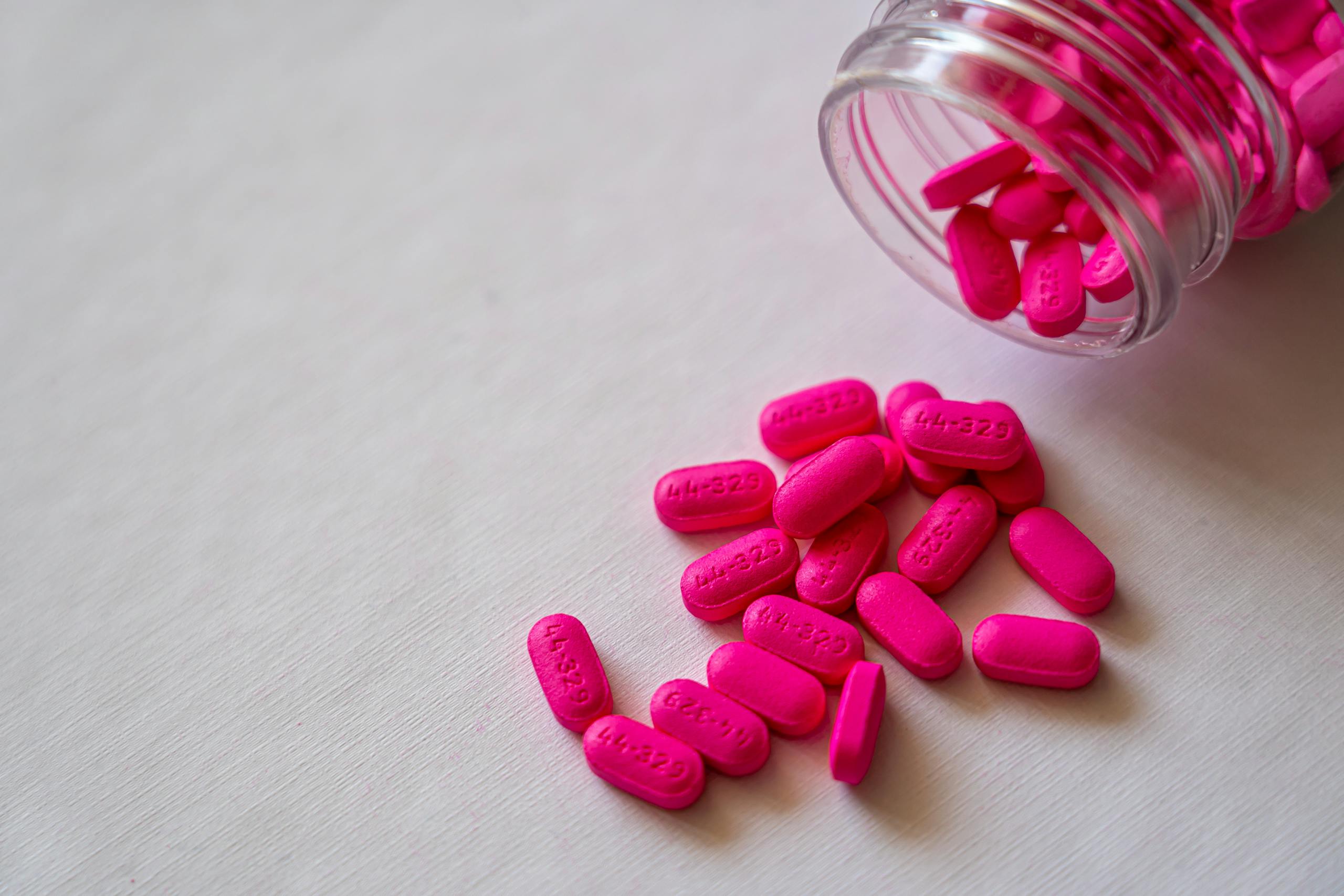 Close-up of vibrant pink pills spilling from a glass jar on a white background, emphasizing healthcare and medication.