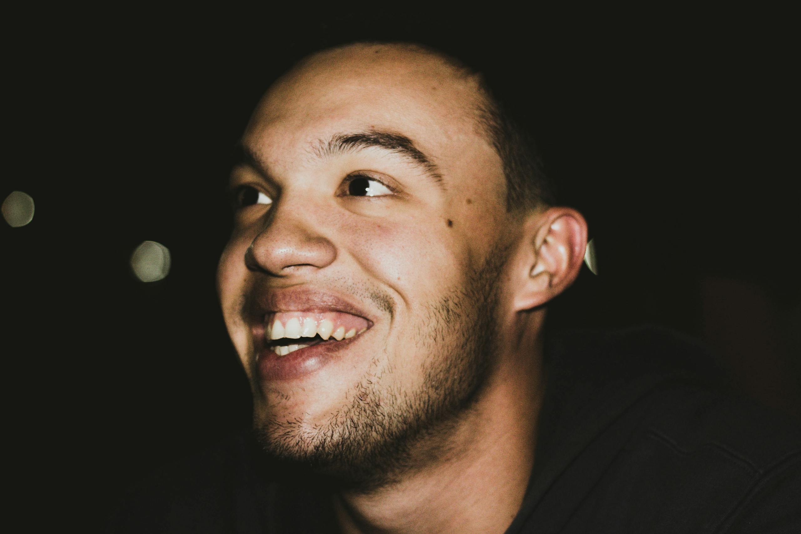 Close-up portrait of a young man with a beard smiling brightly against a dark background.