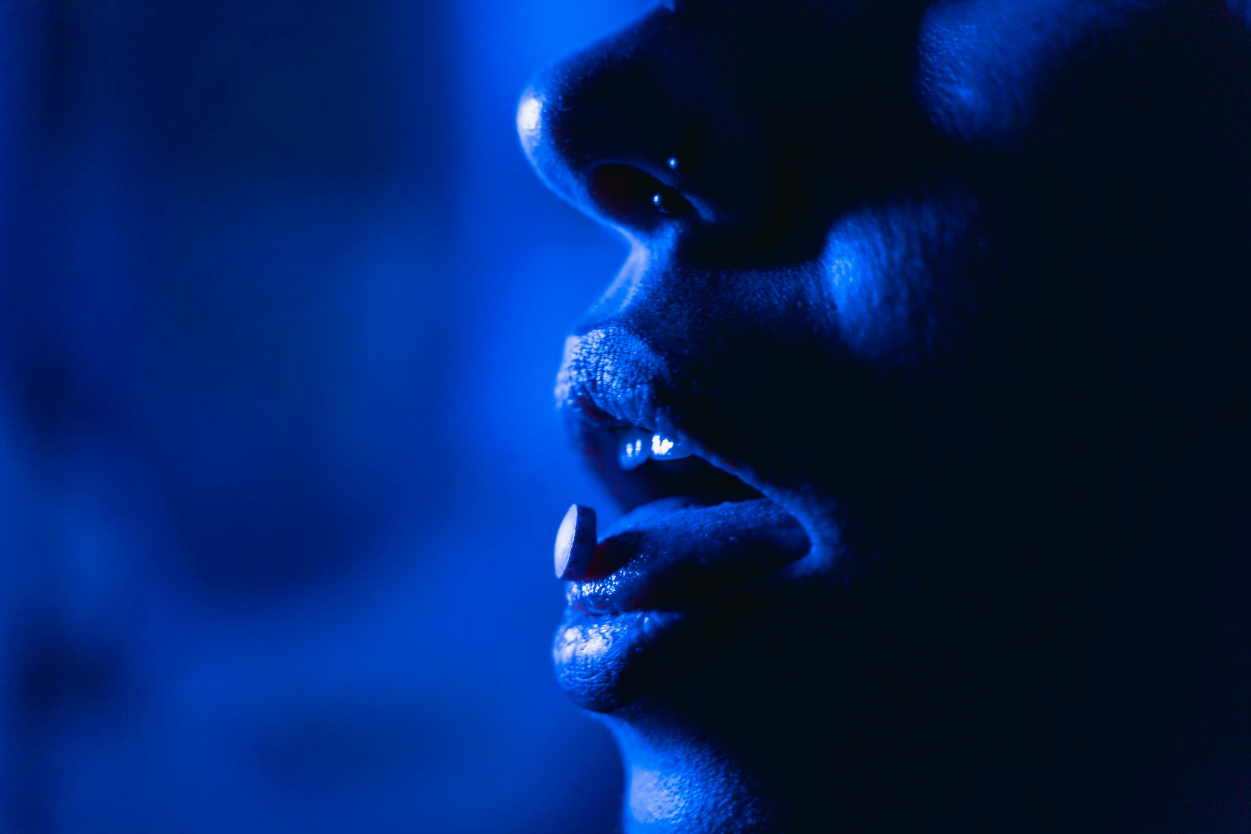 Close-up shot of a person holding a pill in their open mouth in dramatic blue lighting.