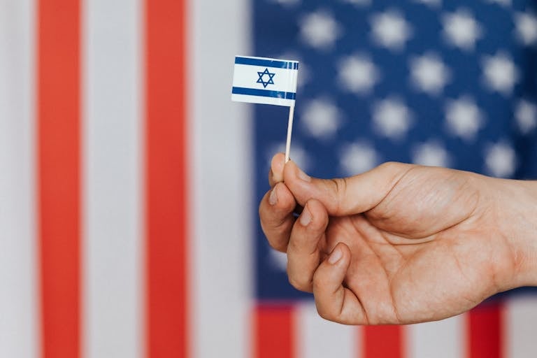Closeup of crop unrecognizable person holding small flag of Israel before huge flag of United States of America on background