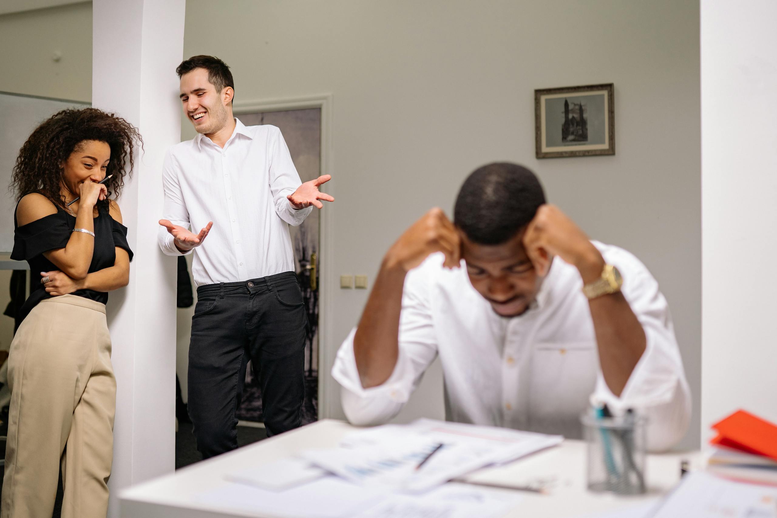 Colleagues sharing a light moment while another looks stressed at his desk in a modern office.