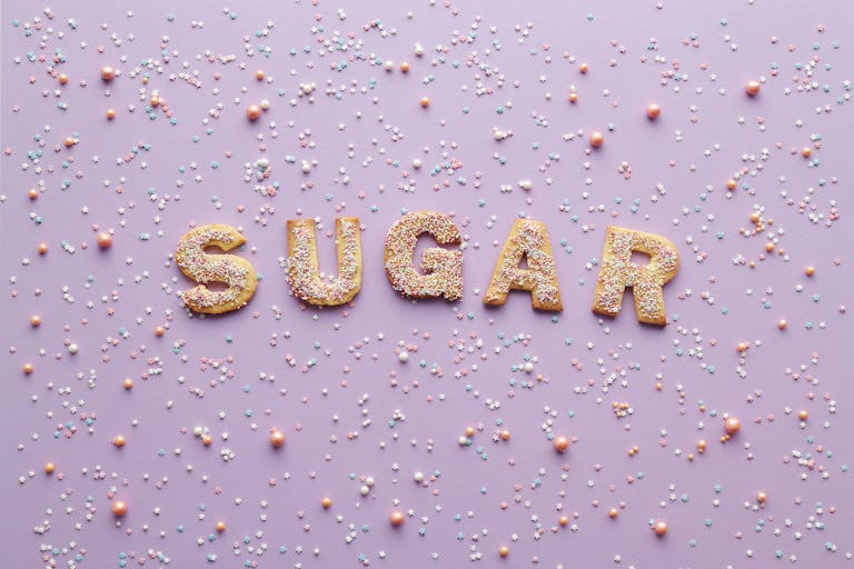 Colorful sprinkle-covered cookies spelling 'sugar' on a pastel purple background, creating a sweet and playful flatlay design.