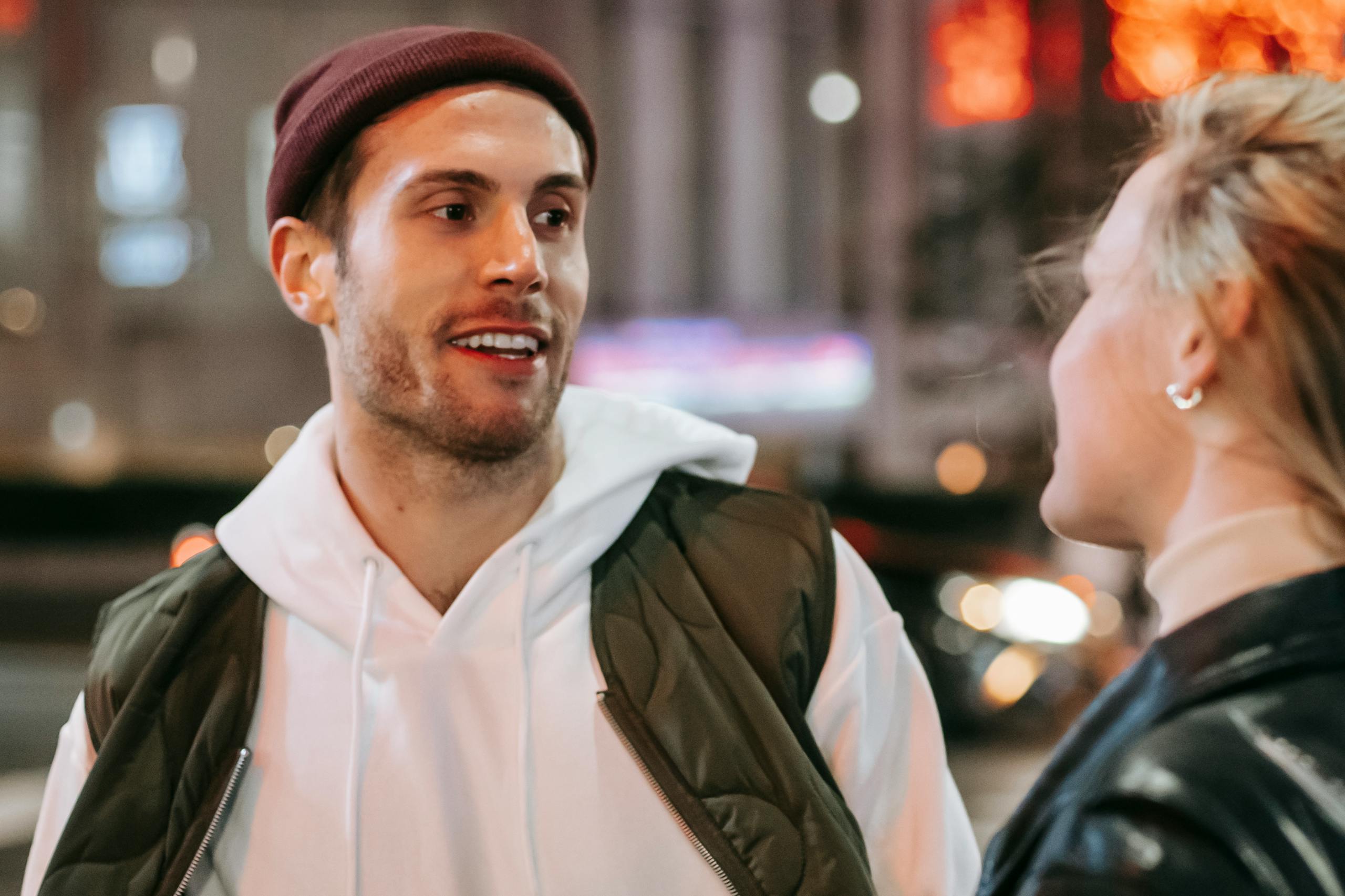 Content man in red hat with unrecognizable woman standing on street with cars and buildings on blurred background at night time