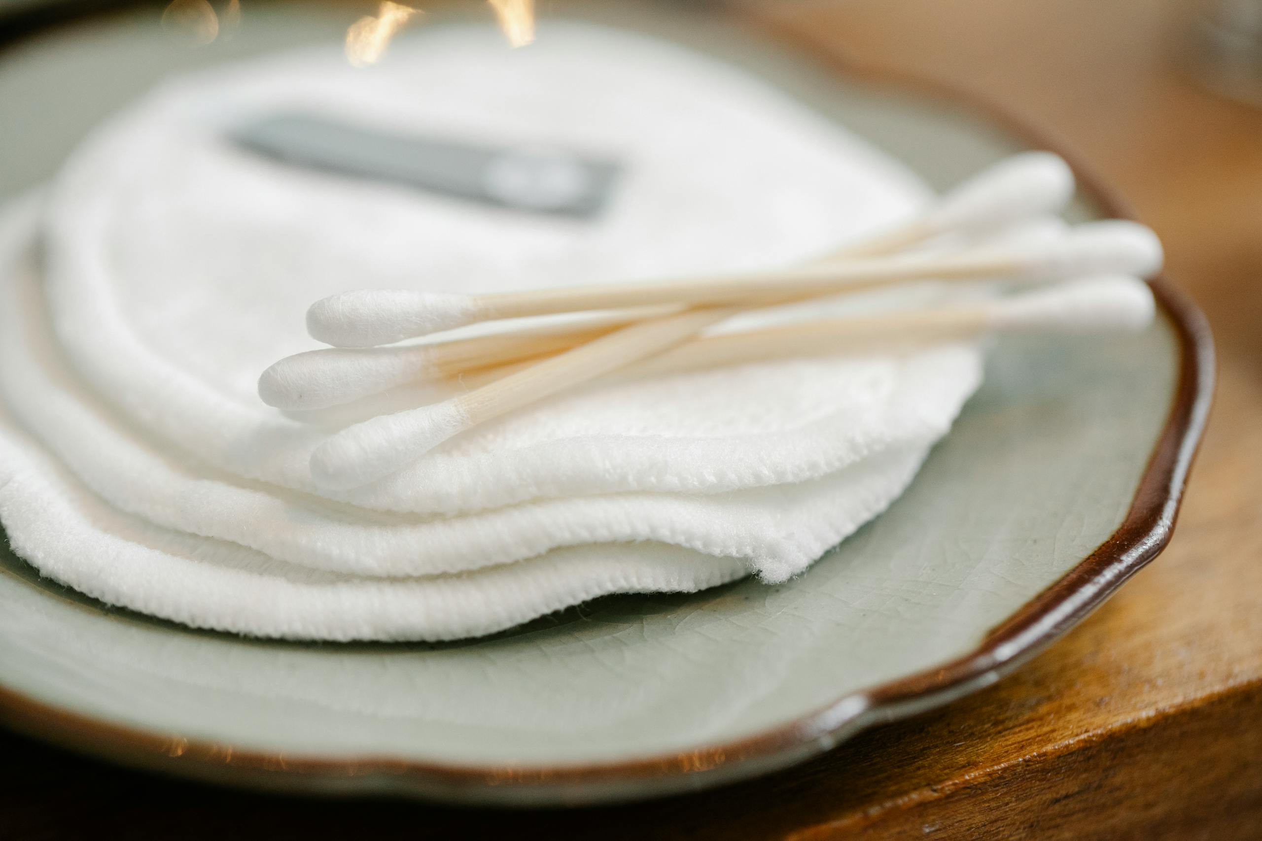Cotton swabs on white round cotton pads for skincare routine placed on plate on wooden table in light room on blurred background