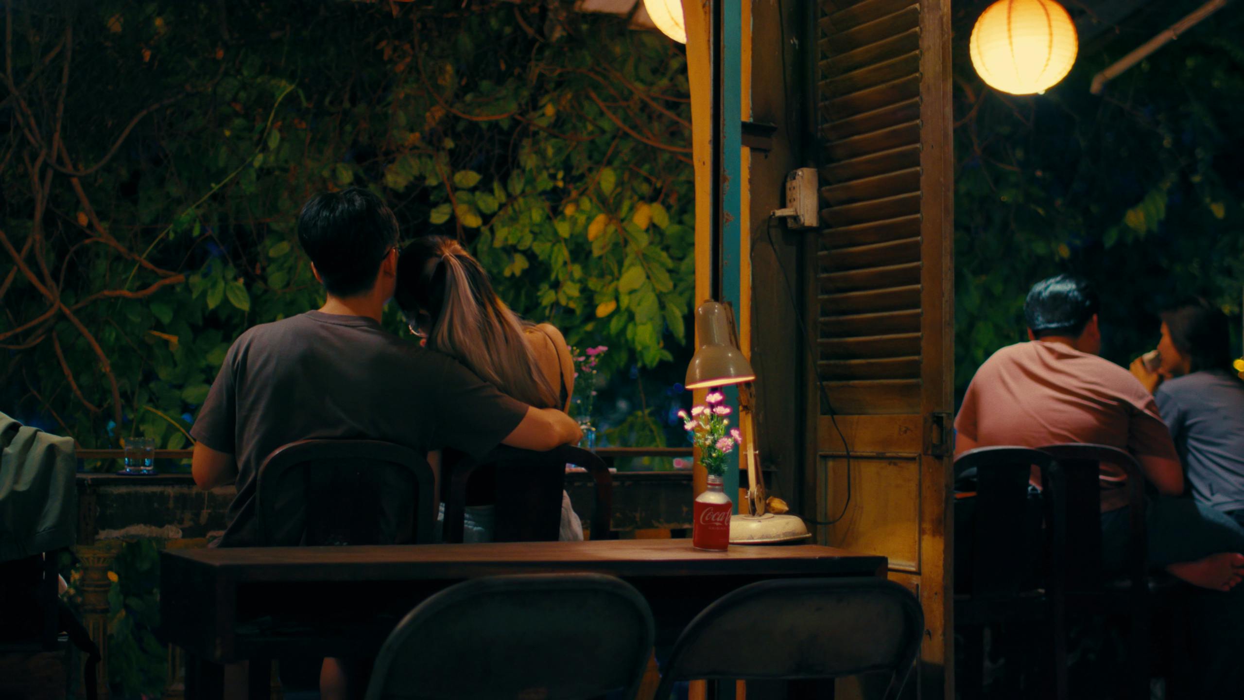 Couple enjoying a romantic evening at a cozy outdoor cafe with warm lighting and lush greenery.