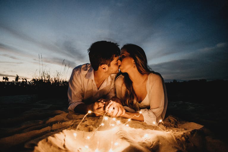 Couple kissing and holding hands while lying near illuminating garland against picturesque sky