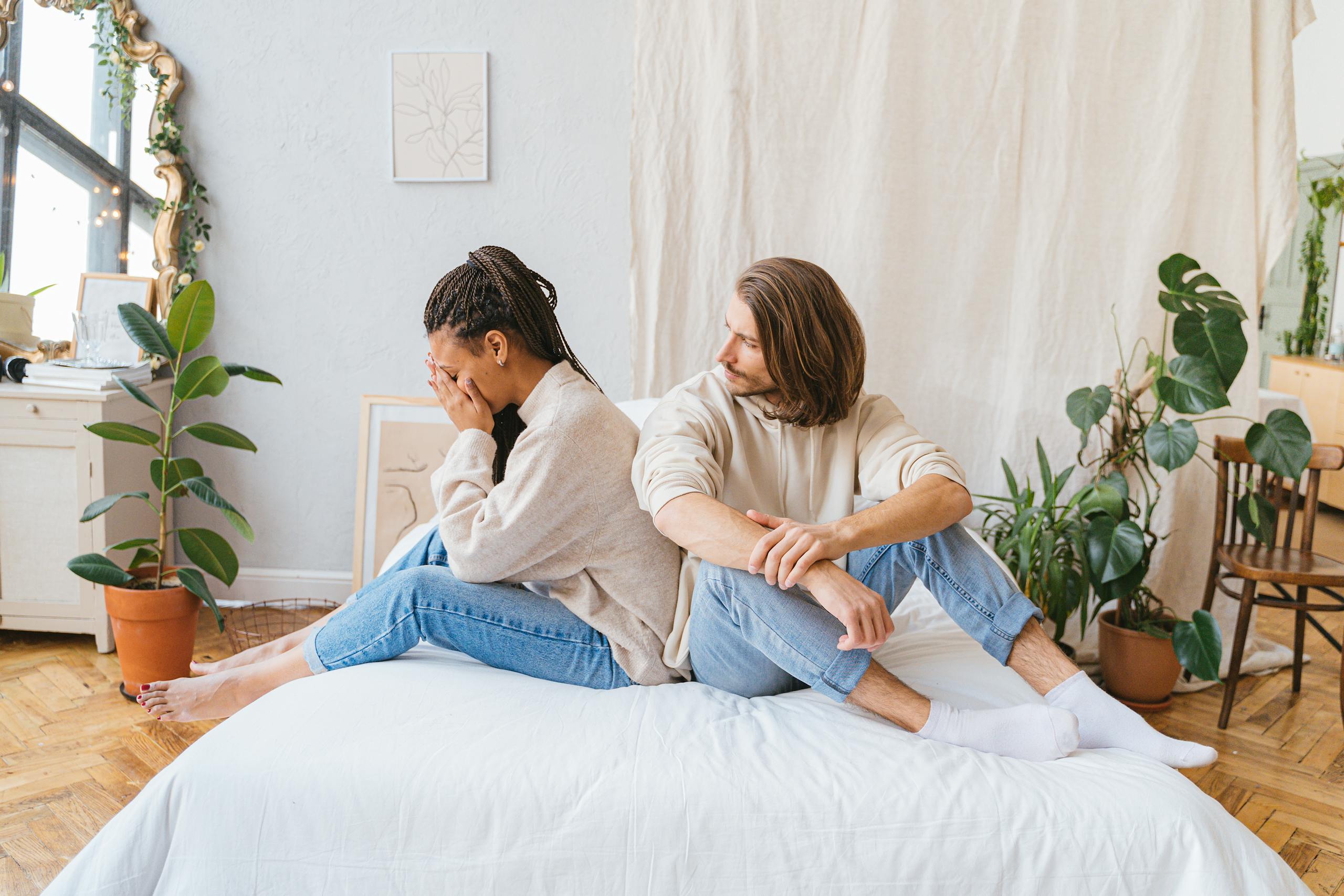 Couple sitting back-to-back on bed, showing relationship conflict in a cozy room.