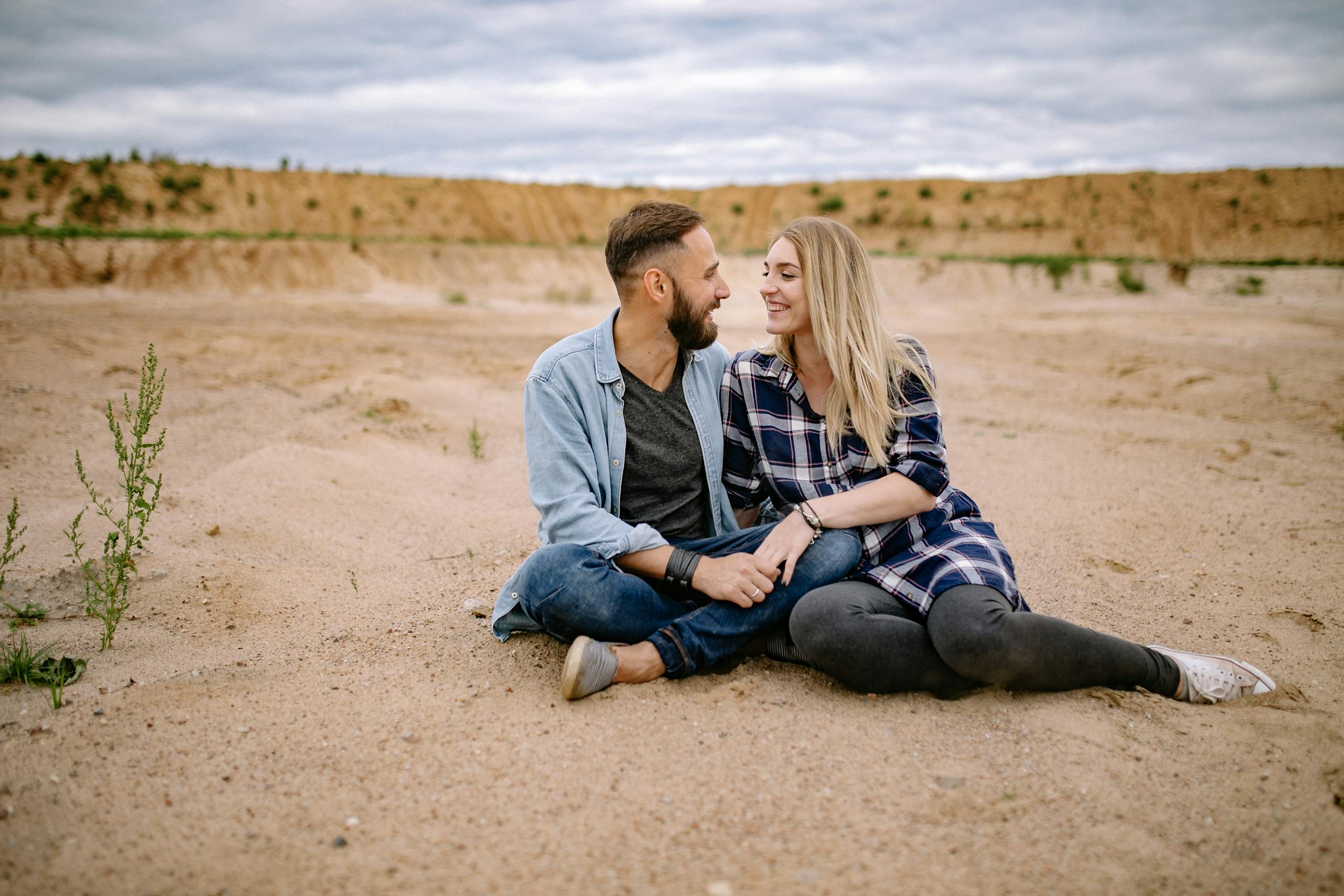 Couple sitting on sand, smiling and holding hands in a serene desert landscape.