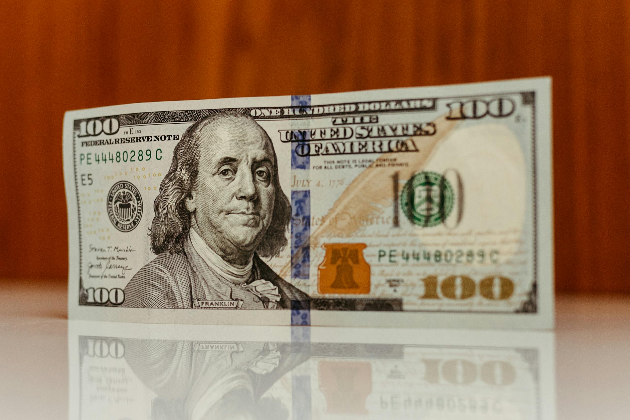 Crisp image of a $100 bill standing and reflecting on a smooth surface, highlighting details.