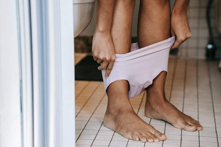 Crop anonymous barefoot ethnic female putting on bikini on tiled floor near toilet at home