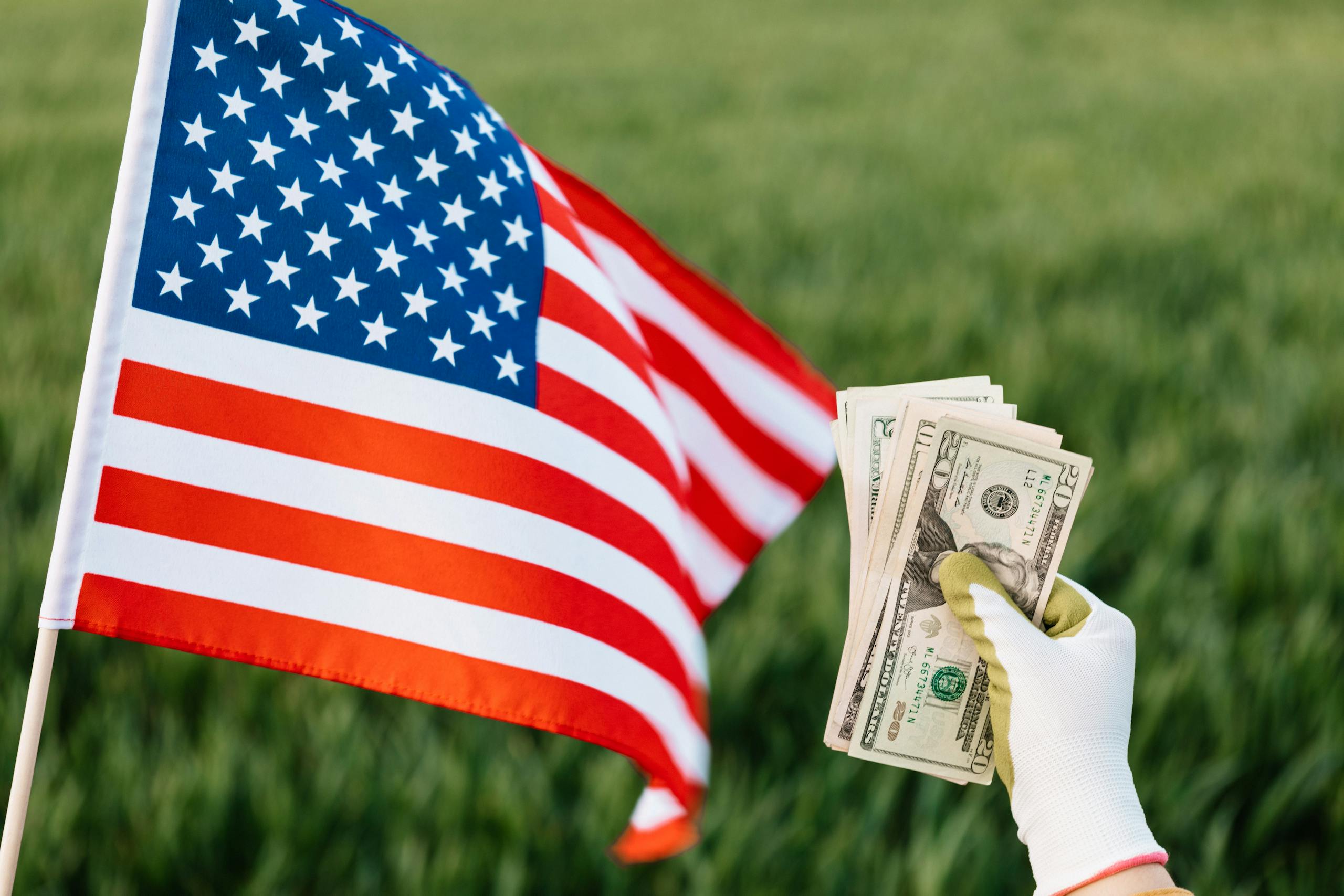 Crop anonymous person in glove with heap of paper money near national flag of United States of America on grass field in daylight