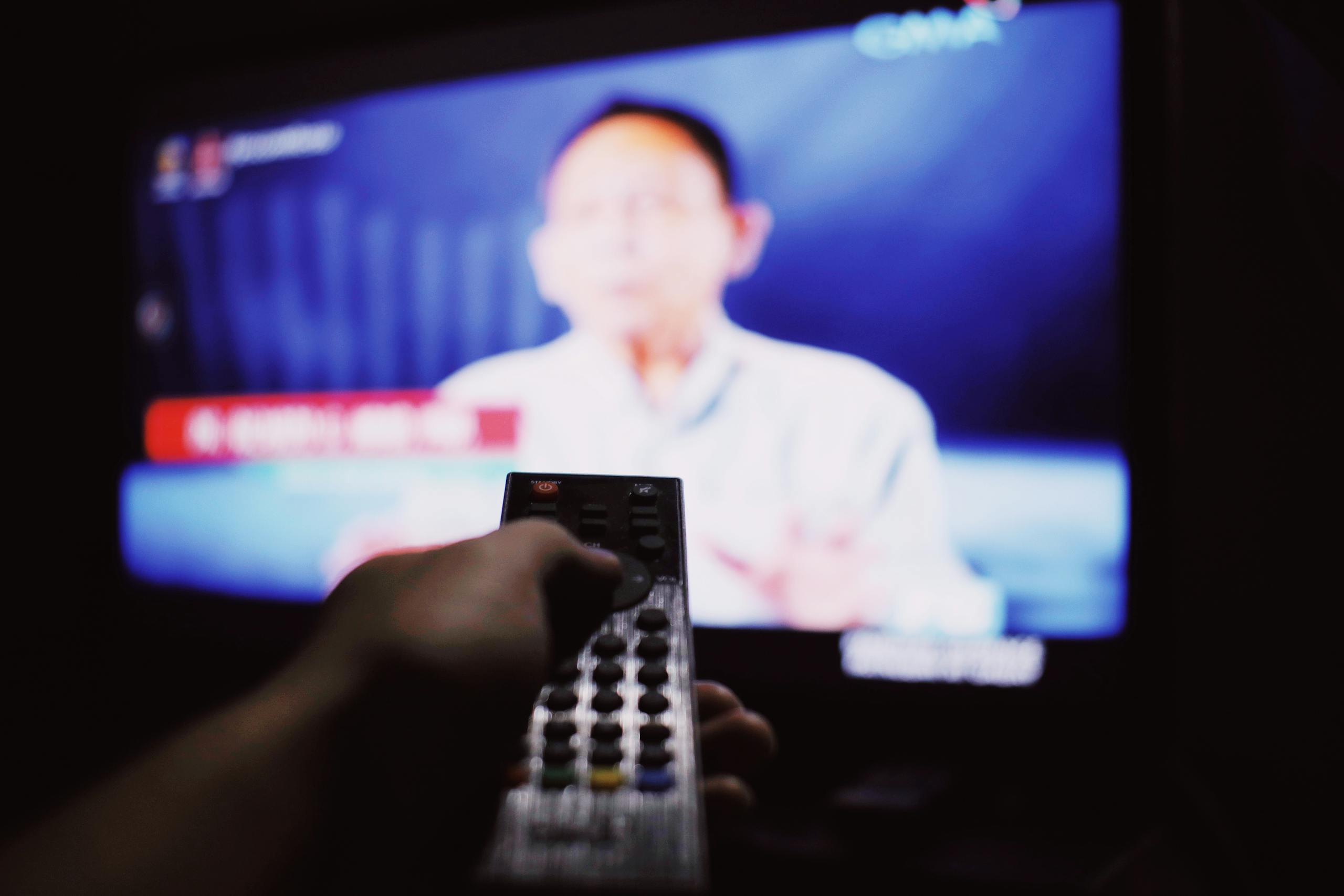 Crop anonymous person with remote controller switching channels while watching TV on blurred background in living room with dim light