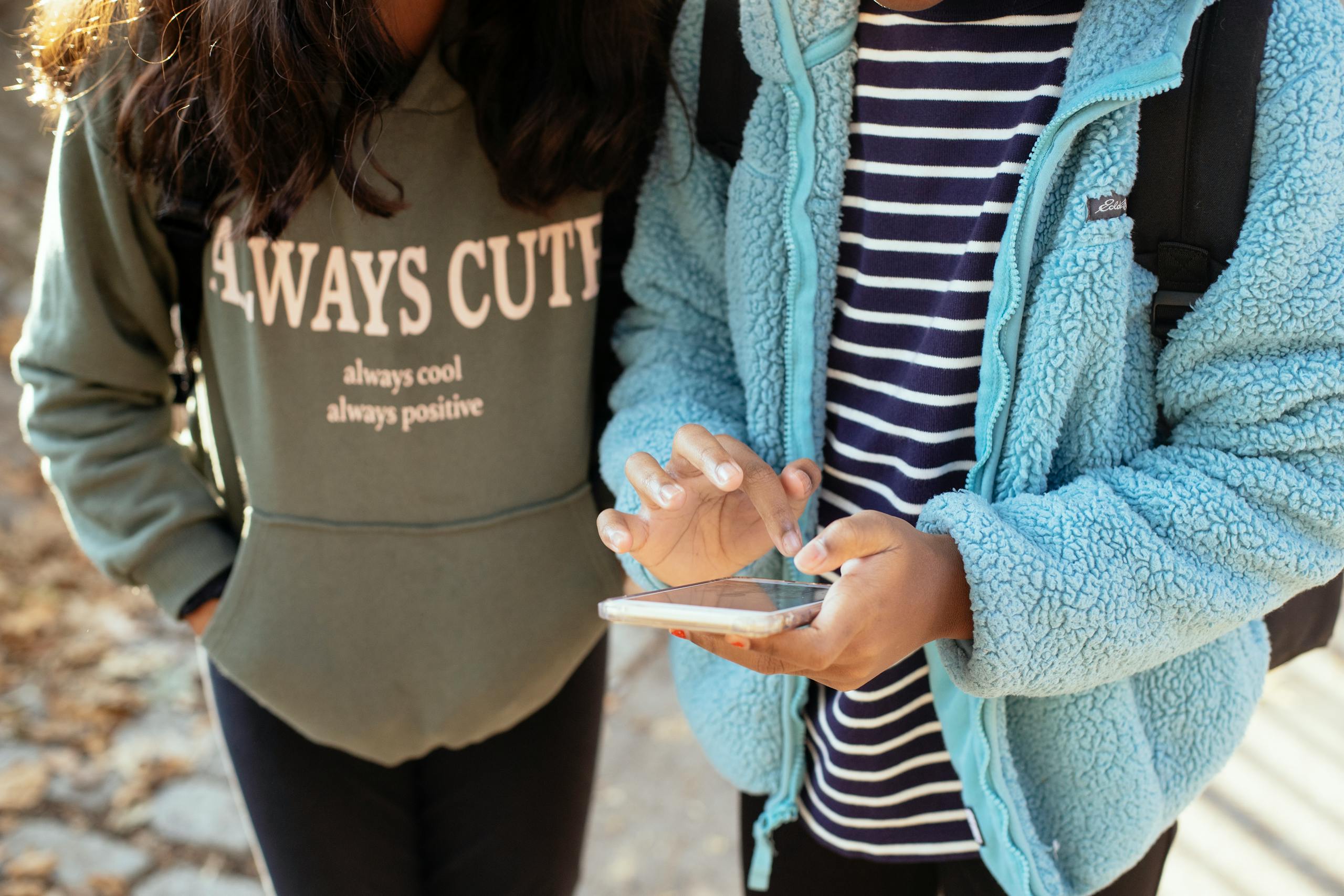 Crop faceless little girls standing on street together and browsing mobile phone while spending weekend