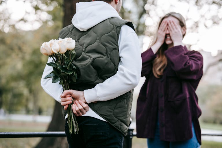 Crop unrecognizable man in warm clothes going to give bouquet of roses as surprise gift to happy girlfriend covering eyes with hands in park