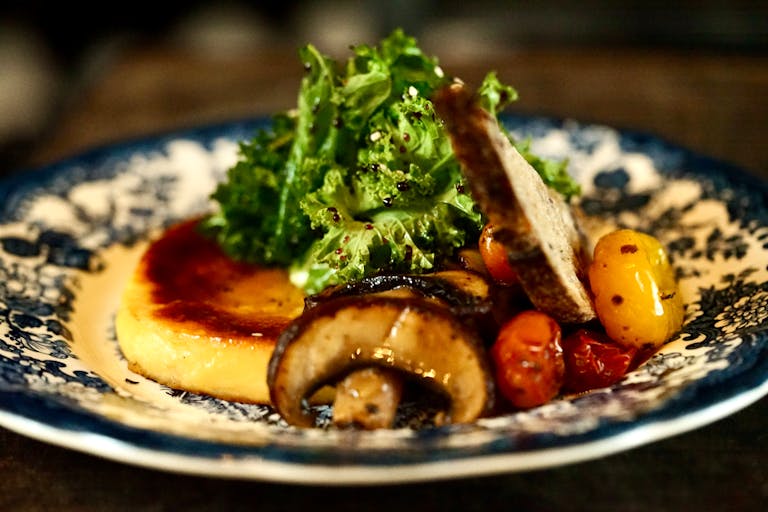 Delicious gourmet dish featuring mushrooms, sauteed greens, polenta, and cherry tomatoes served on a vintage plate.