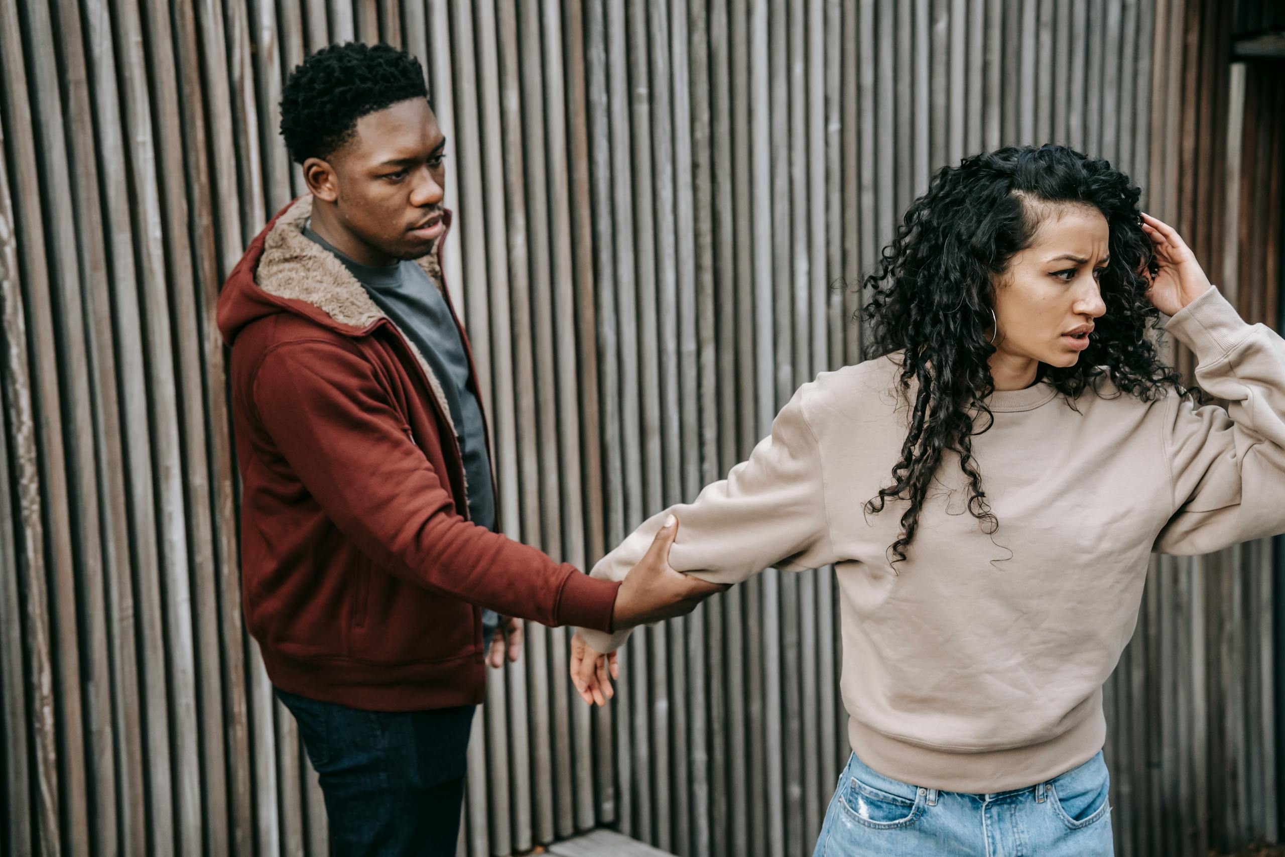 Disappointed African American male trying to stop unhappy ethnic girlfriend while having argument on street near wooden fence during breakup