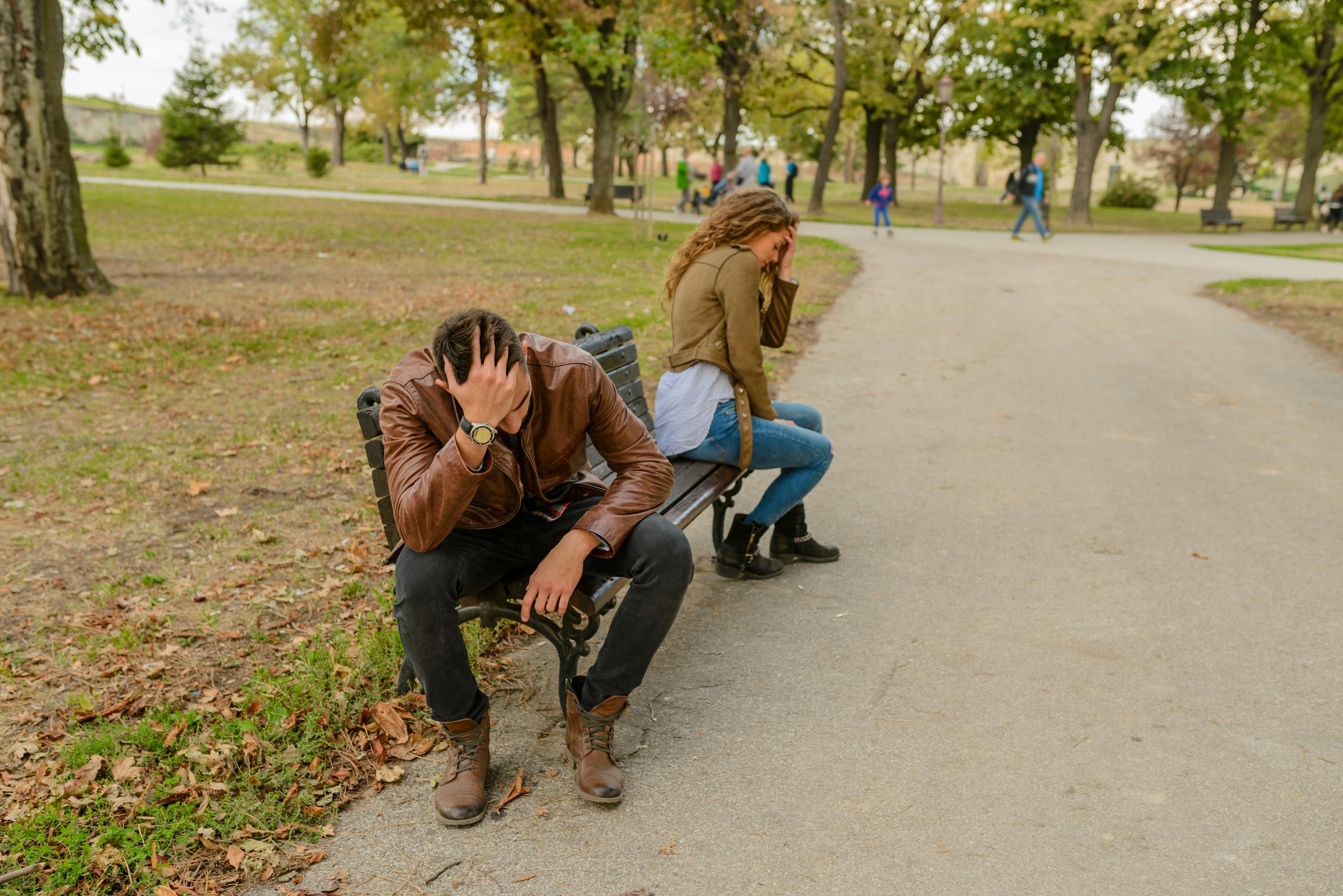 Distraught couple sitting apart on a park bench, facing away from each other, in a public park setting.