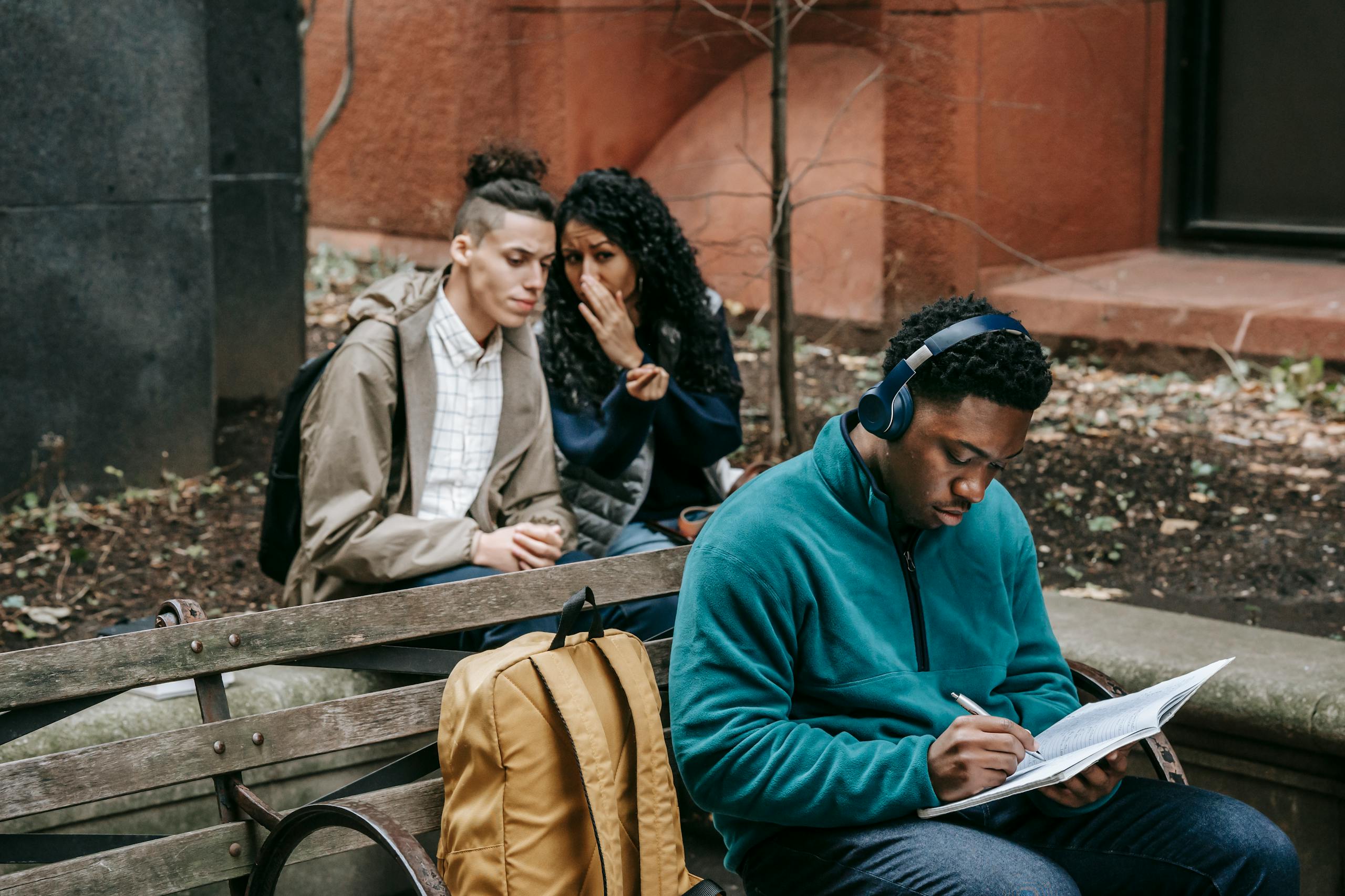 Diverse friends spreading gossip while sitting behind man with headphones and notepad on bench