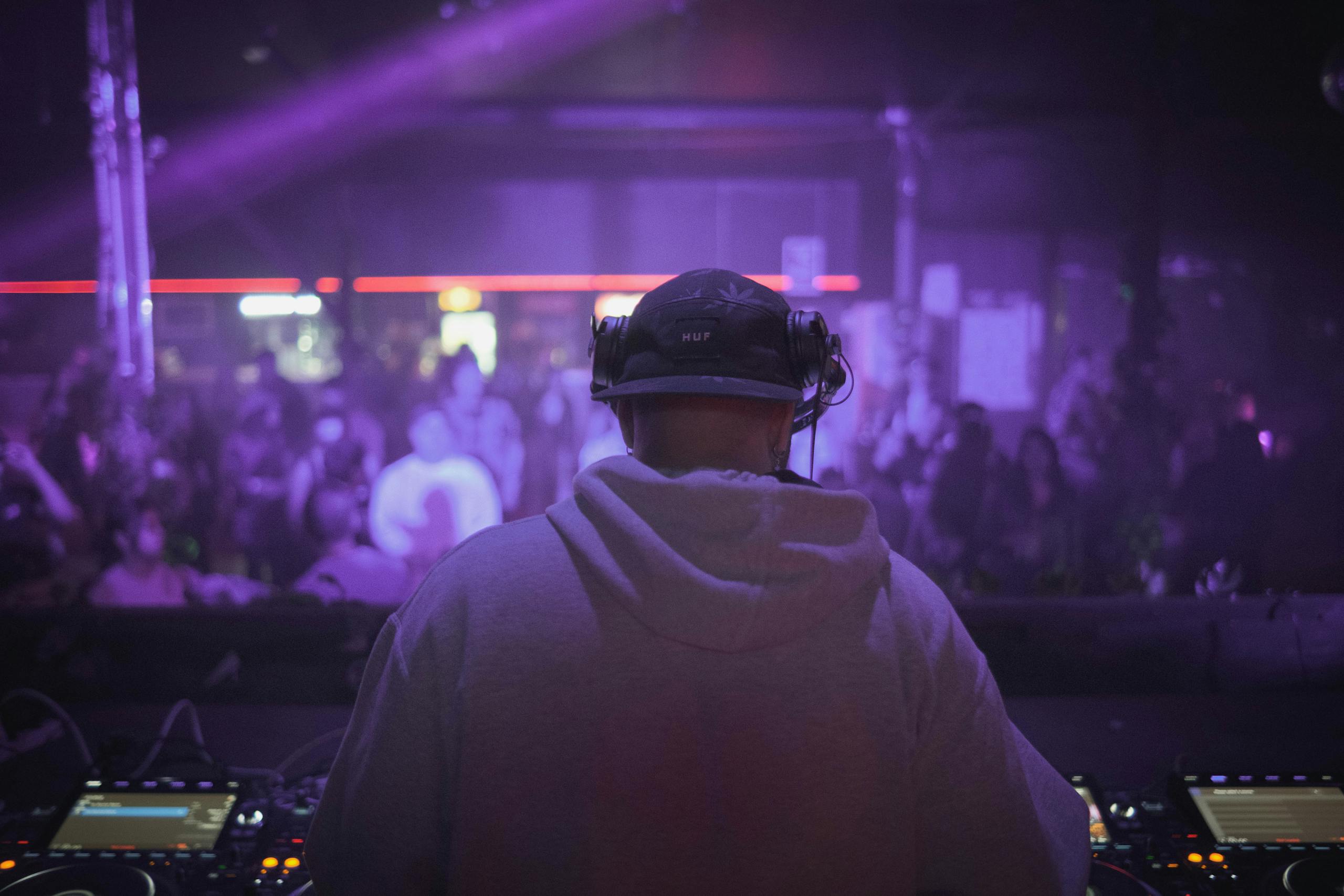 DJ in hoodie playing live set at a vibrant nightclub with purple lighting.