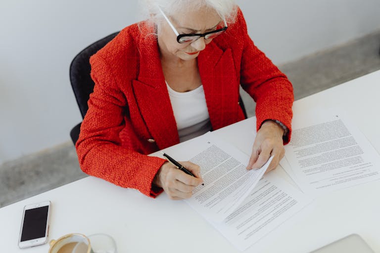 Elderly businesswoman in red jacket signing papers at office desk, showcasing leadership.