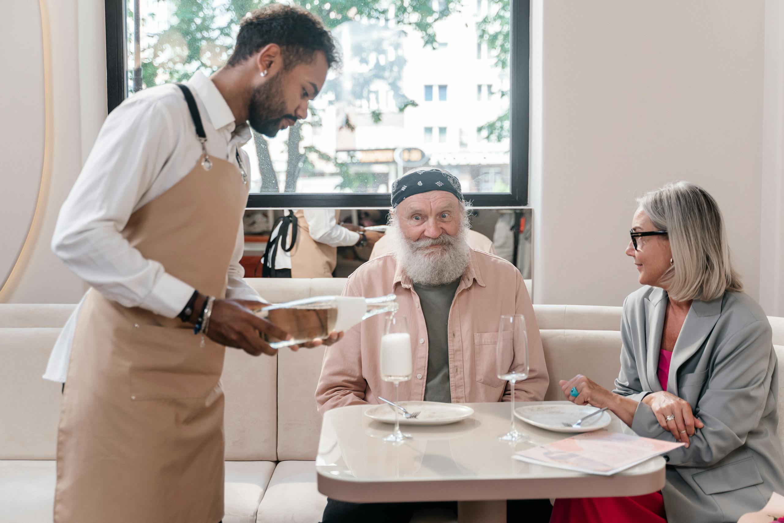 Elderly couple being served champagne by a waiter in a restaurant setting.