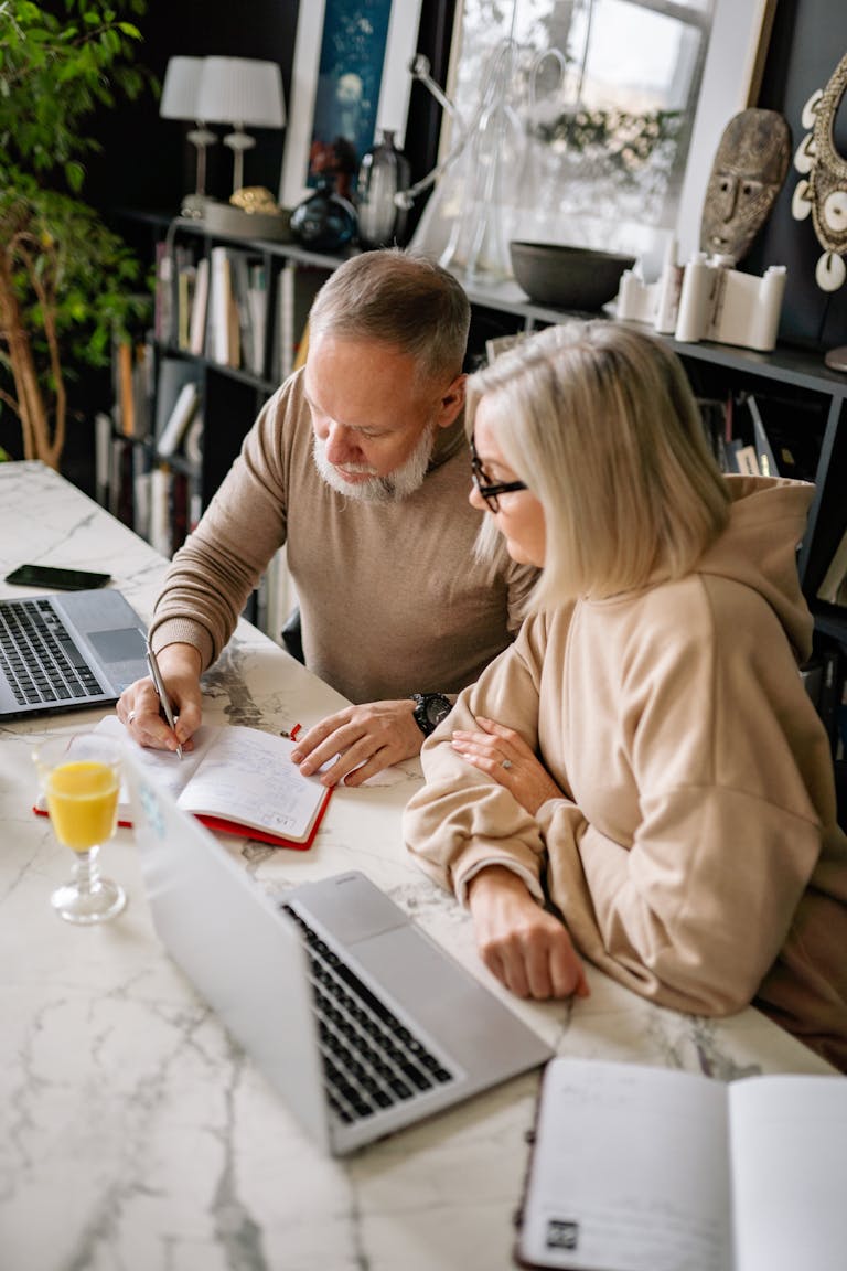 Elderly couple working together at home using laptops and a notebook in a cozy setting.