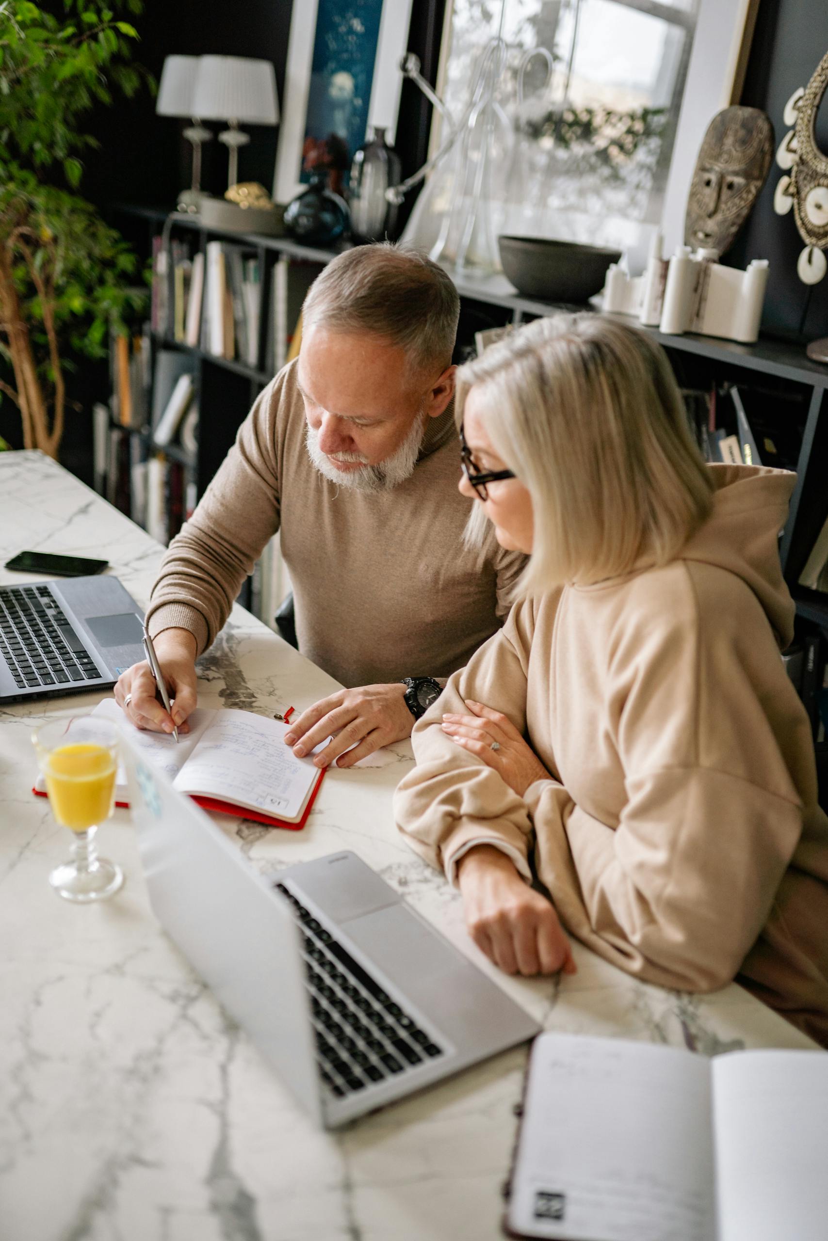 Elderly couple working together at home using laptops and a notebook in a cozy setting.