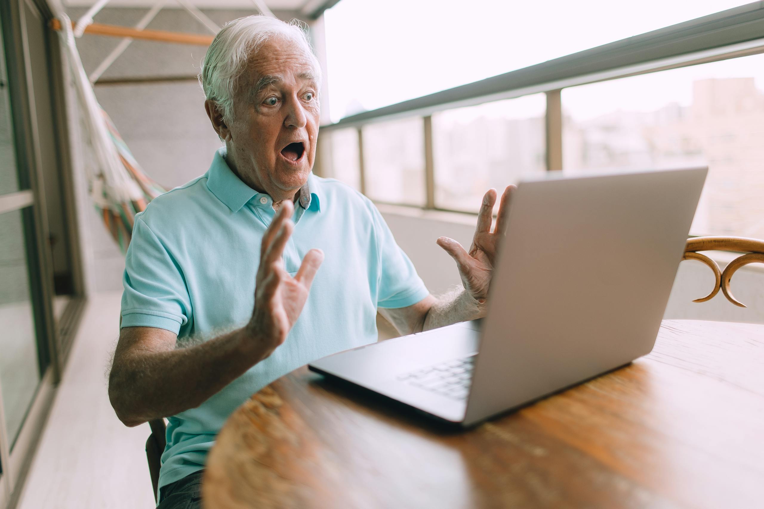 Elderly man looking shocked while using a laptop on a balcony, depicting excitement or surprise.