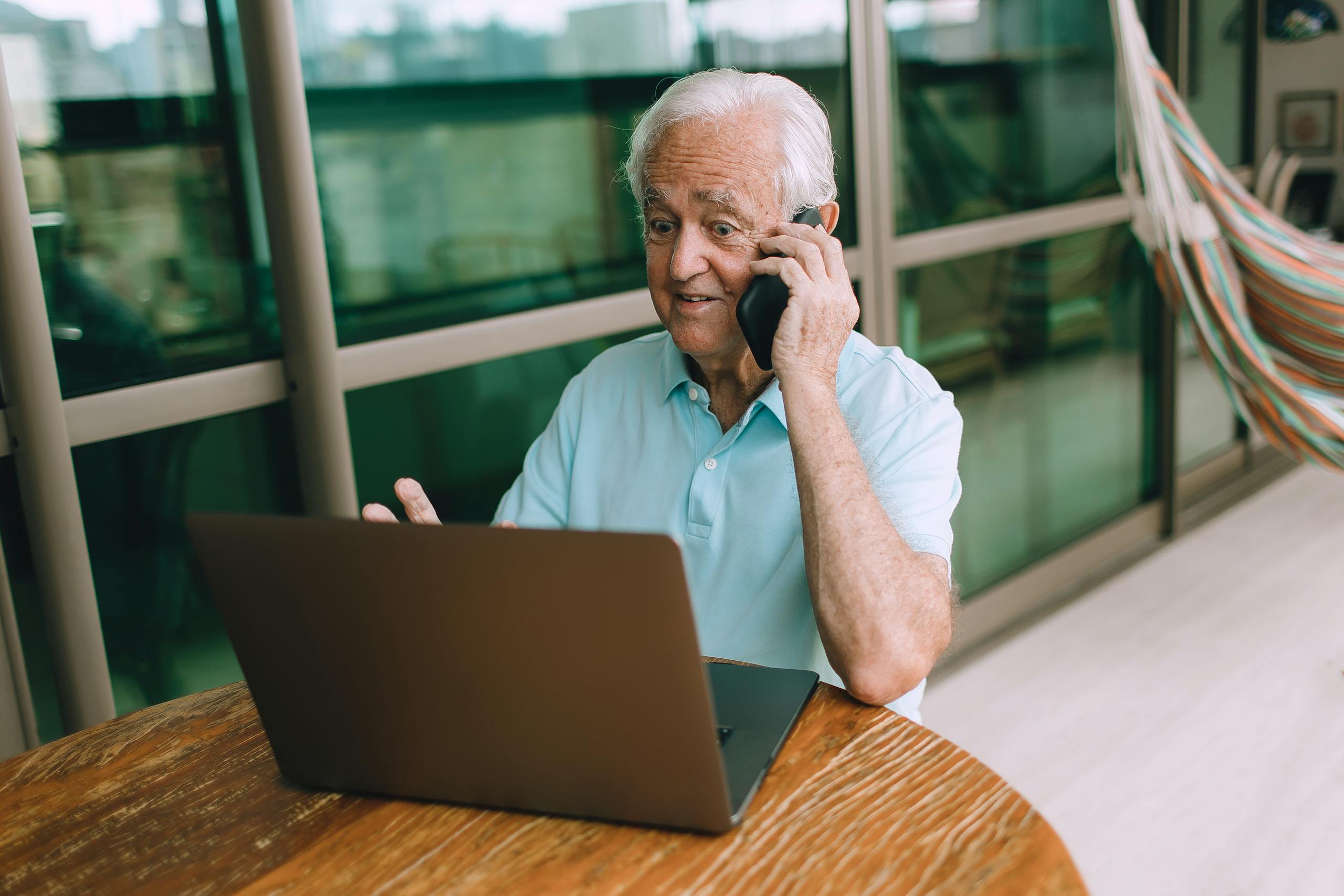 Elderly man multitasking on laptop and phone indoors, focused on technology.