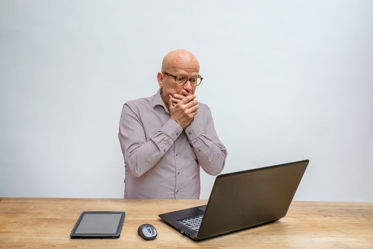 Elderly man reacts in surprise to laptop screen at wooden desk with tablet and mouse.