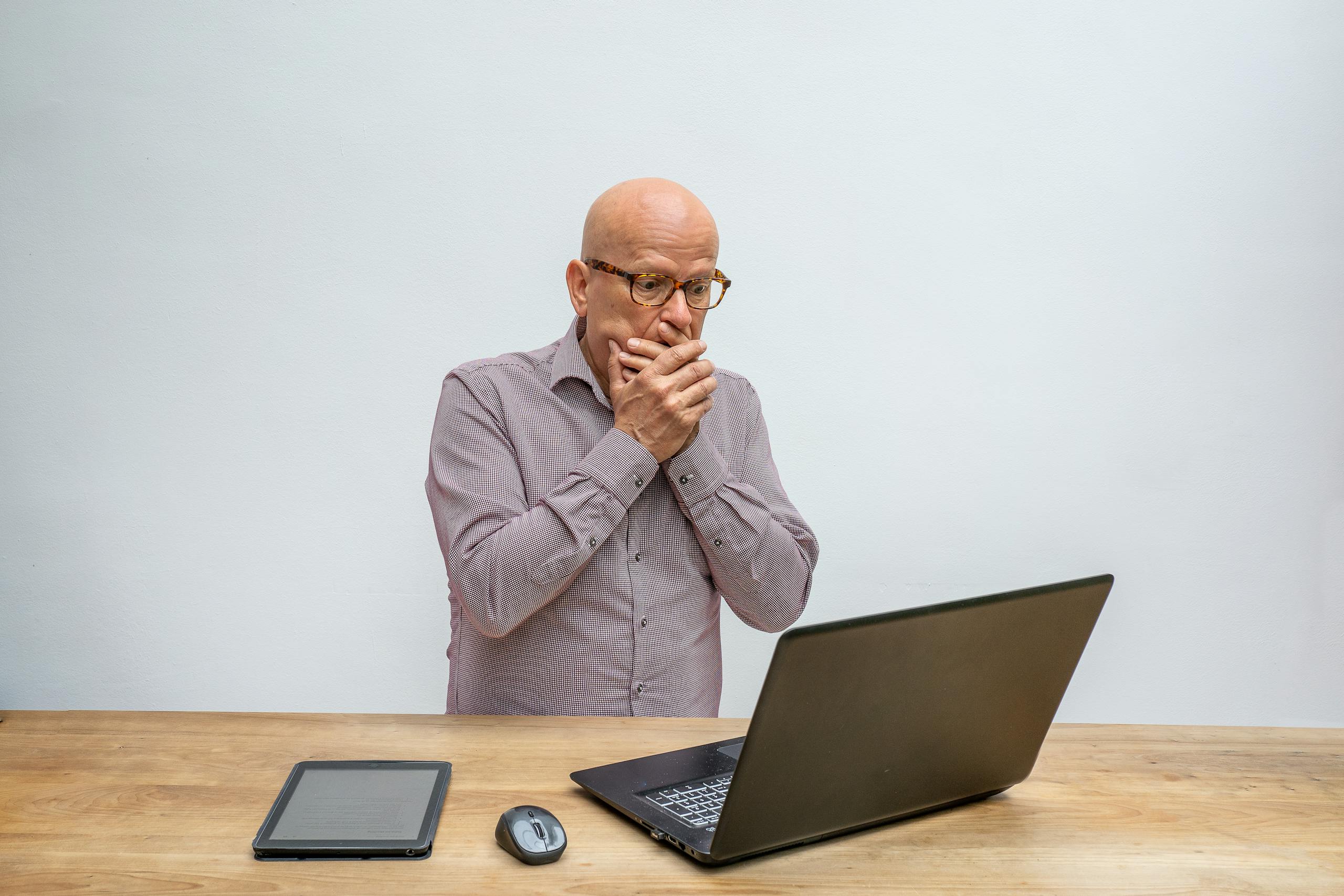 Elderly man reacts in surprise to laptop screen at wooden desk with tablet and mouse.