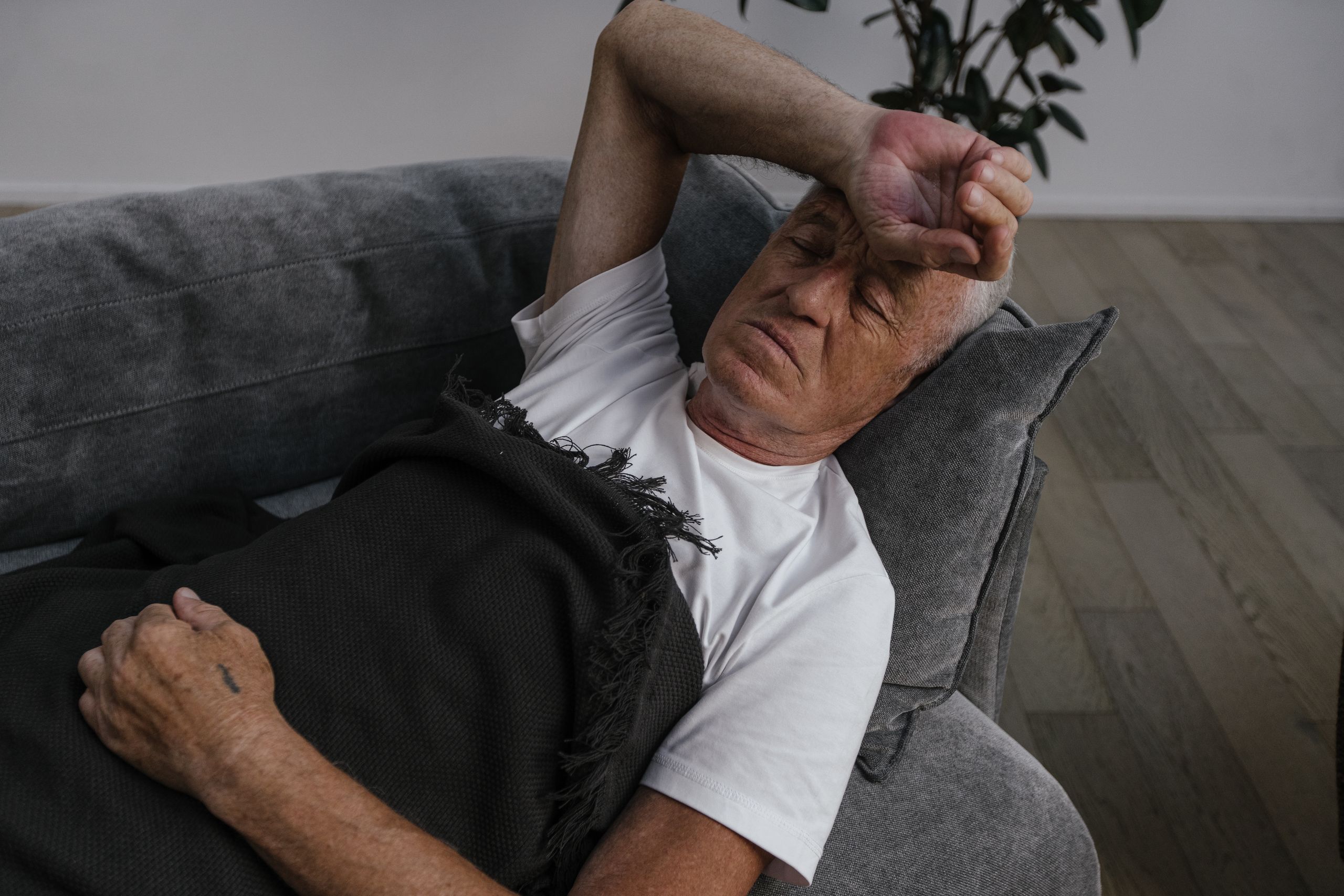Elderly man relaxing on a sofa, hand on forehead, white shirt and blanket.