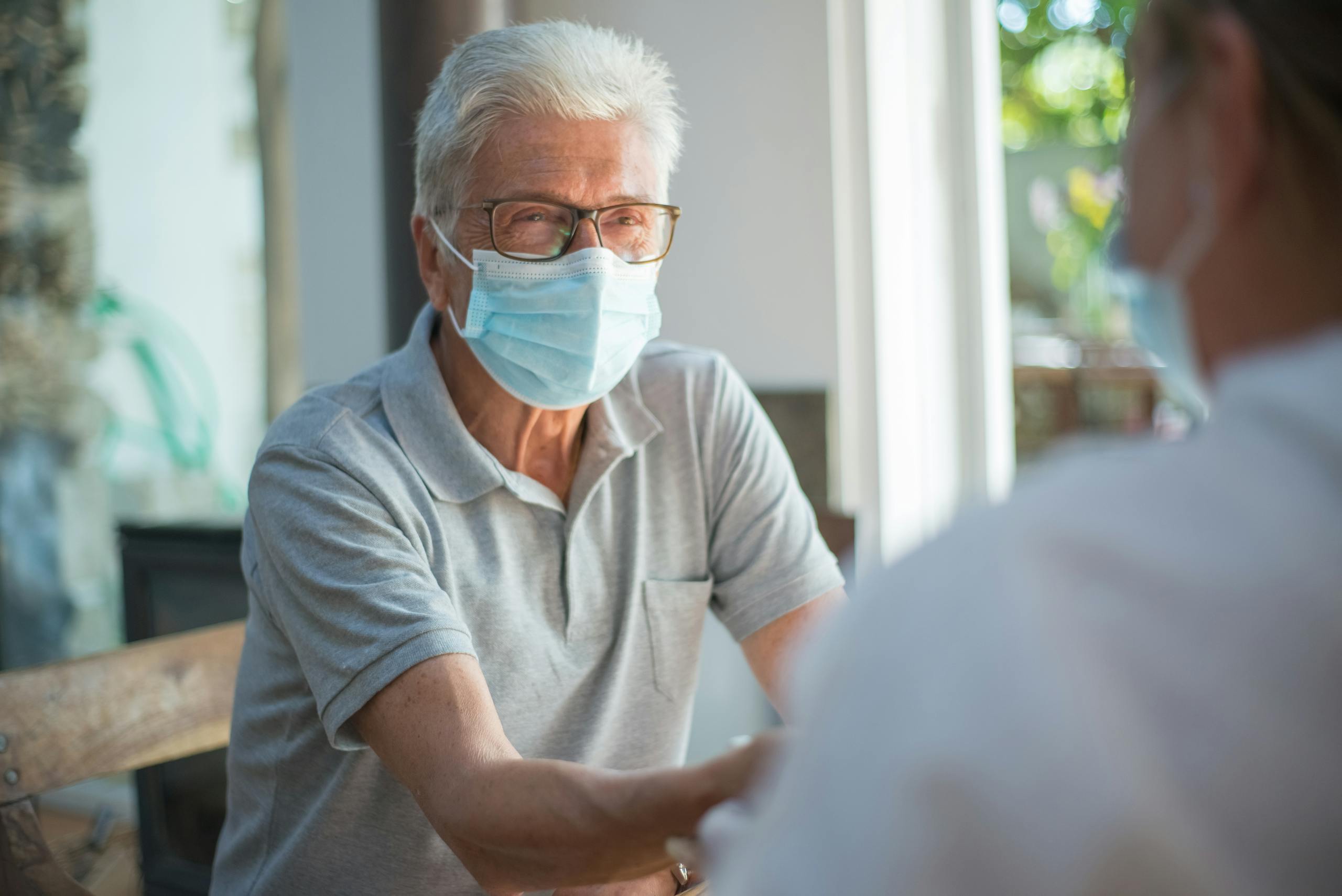 Elderly man sitting indoors wearing a mask and eyeglasses during a discussion. Bright and relaxed atmosphere.