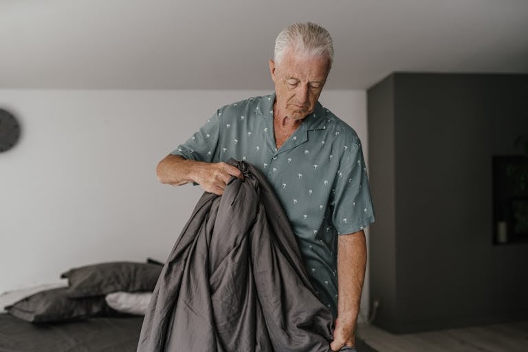Elderly man with gray hair adjusting a bed. Indoor lighting sets a calm tone.