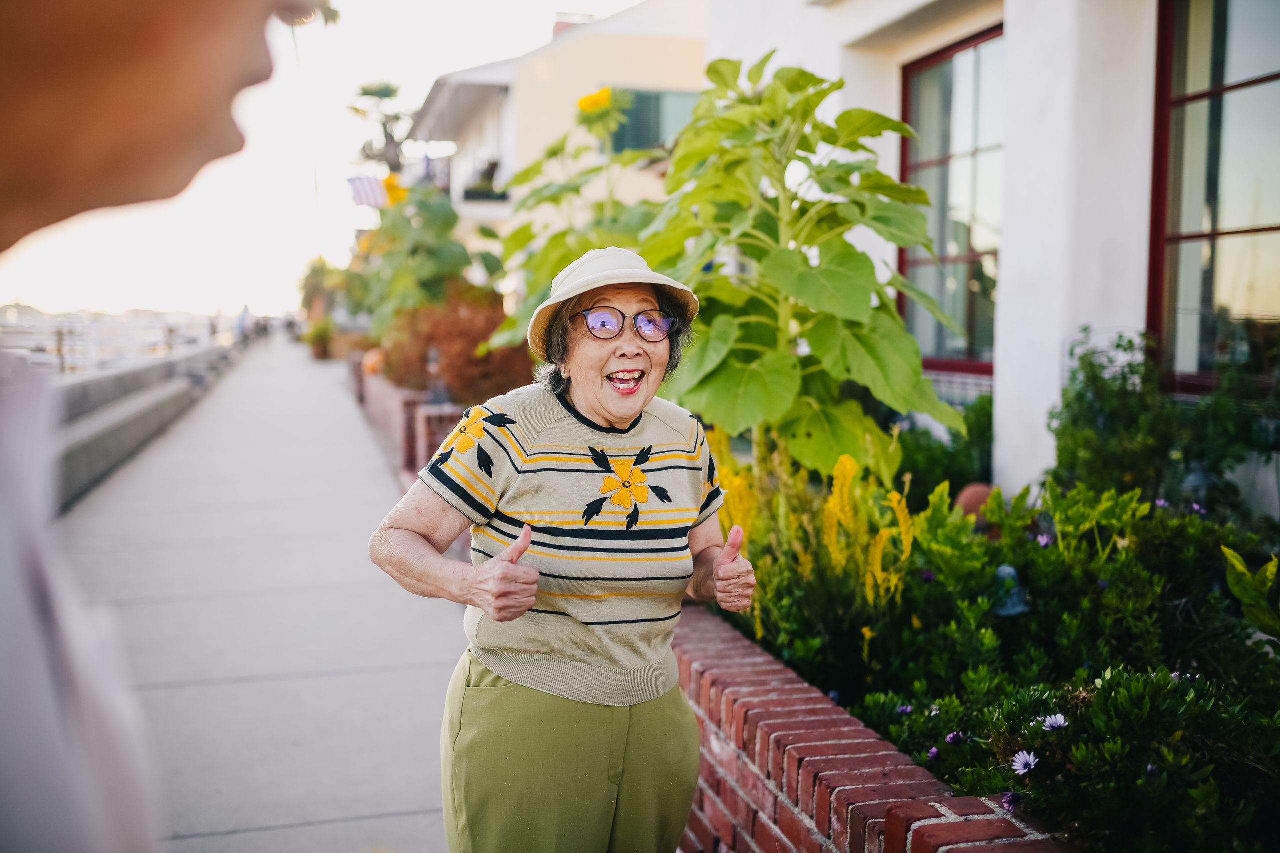 Elderly woman with thumbs up, happy in a garden setting during the day.
