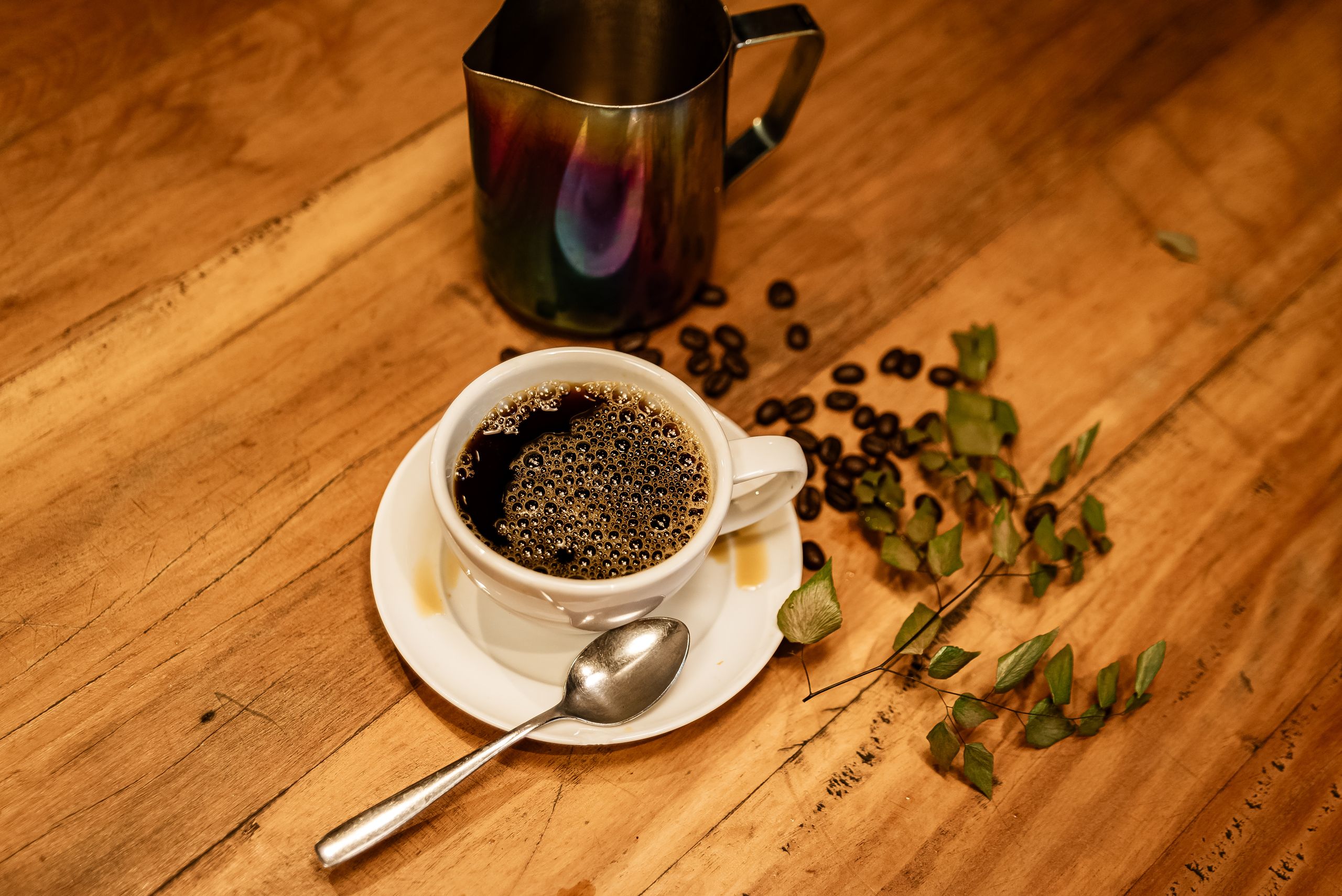 Elegant coffee scene with a coffee cup, roasted beans, and a metal jug on a wooden table.